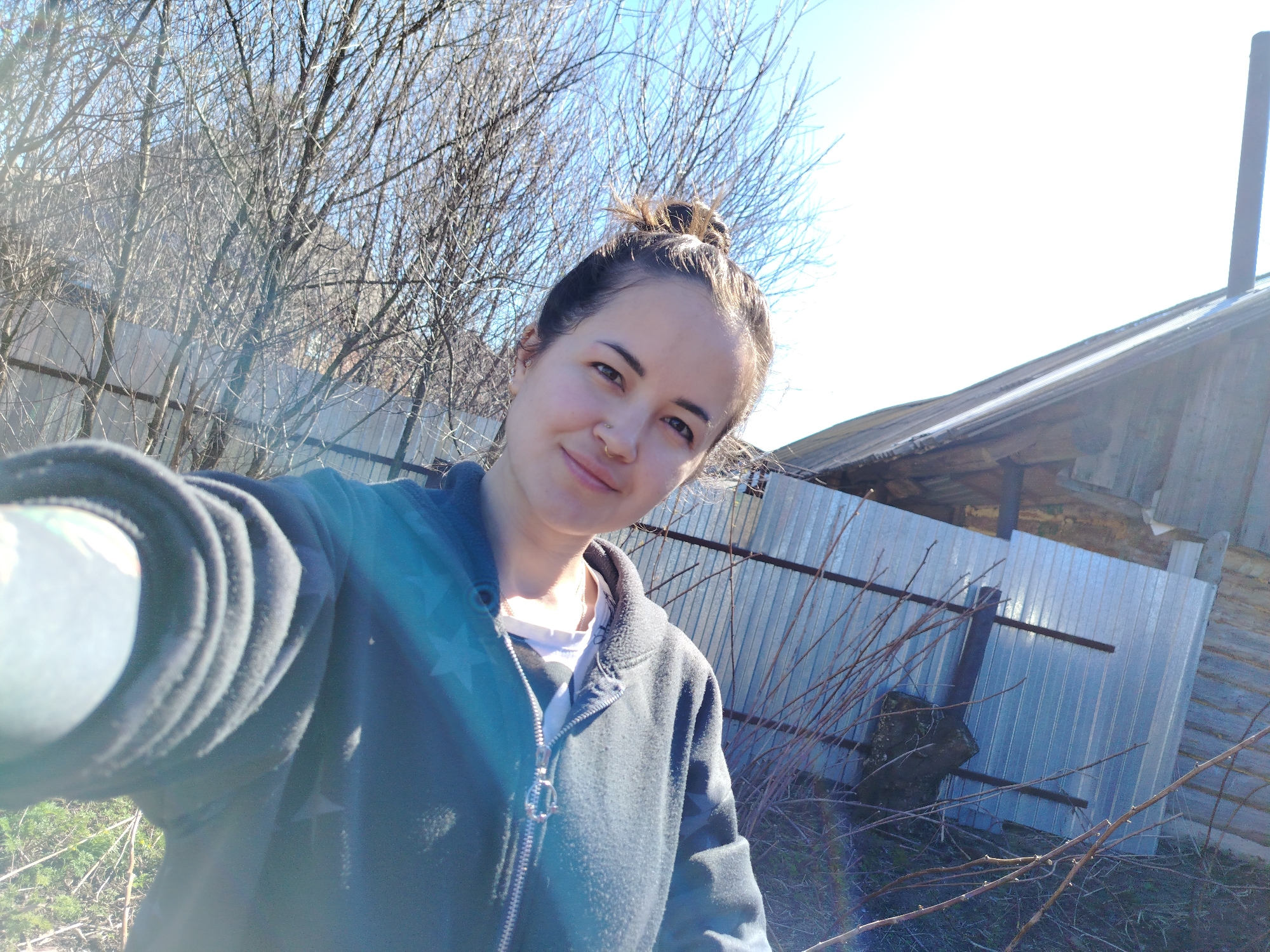 A woman takes a selfie outdoors in Гожан, Russia. She has brown hair in a bun, a light complexion, and wears a zippered gray jacket over a white top. Behind her is a corrugated metal fence, bare trees and branches, and a wooden building with a chimney. The sky is pale blue. Sunlight is visible in the lower left of the frame.