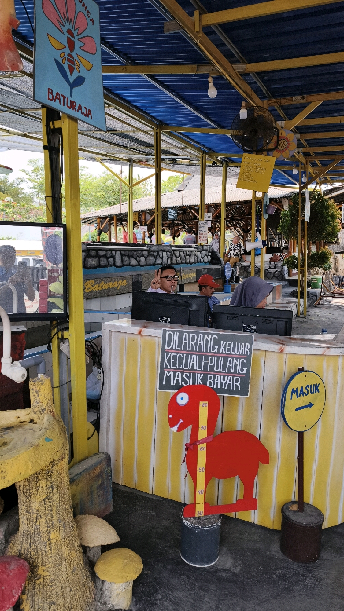 The image depicts an outdoor setting, likely a tourist attraction or a park, in Air Paoh, Indonesia. In the foreground, a yellow and white striped counter is visible, with two individuals working behind it, each looking at a computer monitor. The person on the left, wearing glasses, is giving a peace sign with their right hand. A sign above the counter reads "DILARANG KELUAR KECUALI PULANG MASUK BAYAR" which translates to "DO NOT GO OUT UNLESS GOING HOME, ENTER PAY." To the right of the counter, a red cutout figure resembling a dinosaur with a measuring scale on its body is placed on a black container. A yellow circular sign with an arrow pointing to the right and the word "MASUK" (Enter) is also present.
In the background, a blue roof structure supported by yellow beams covers the area. A sign with a drawing of a flower and the text "BATURAJA" is hanging from a yellow pole. There are other signs and structures suggesting a commercial or recreational establishment. The environment appears to be a warm, possibly tropical, climate with trees and vegetation visible in the distance. The overall scene conveys a lively and active atmosphere.
