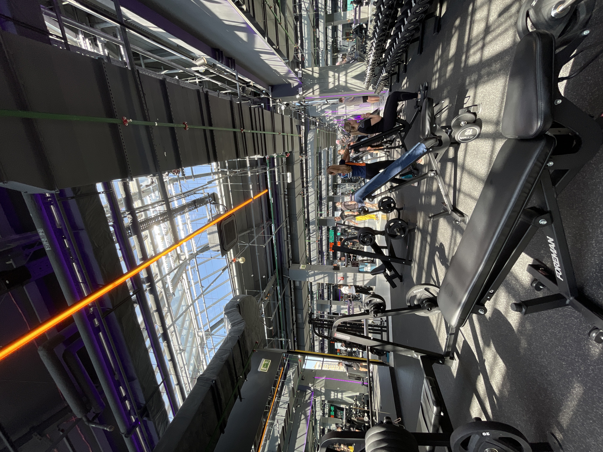 The image depicts the interior of a gym, likely in Kazan, Russia. The space features a high ceiling with visible support structures and a glass roof allowing natural light. Various exercise machines and free weights are arranged throughout the gym, including weight benches, dumbbells, and cable machines. Several individuals are actively engaged in exercise, some using the equipment. The lighting is a combination of natural light from the roof and artificial lighting. The floor is a dark, possibly rubberized material commonly found in gyms. An orange beam of light crosses the top left area of the image.