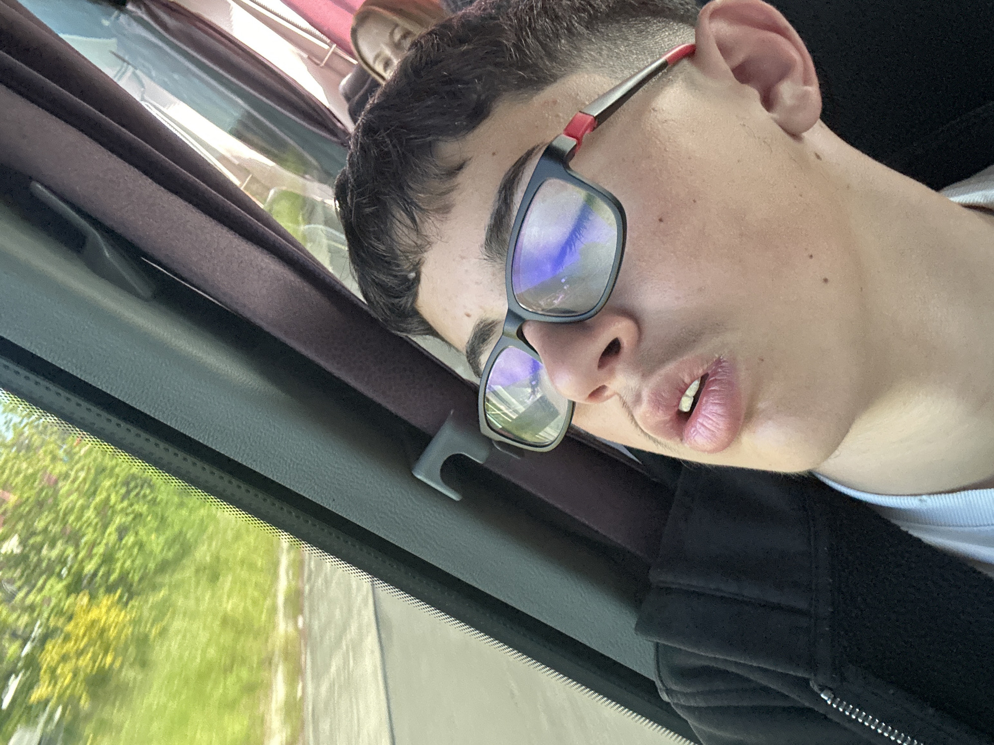 A person with short dark hair and wearing glasses is sleeping in a vehicle. The person is wearing a black jacket over a white shirt. The vehicle is moving, as evidenced by the blurred scenery visible through the window. Another person's face is visible in the background. The location is suggested to be in the Municipal Unit of Kato Olympos, Greece, but there is no visual information in the image to confirm this.