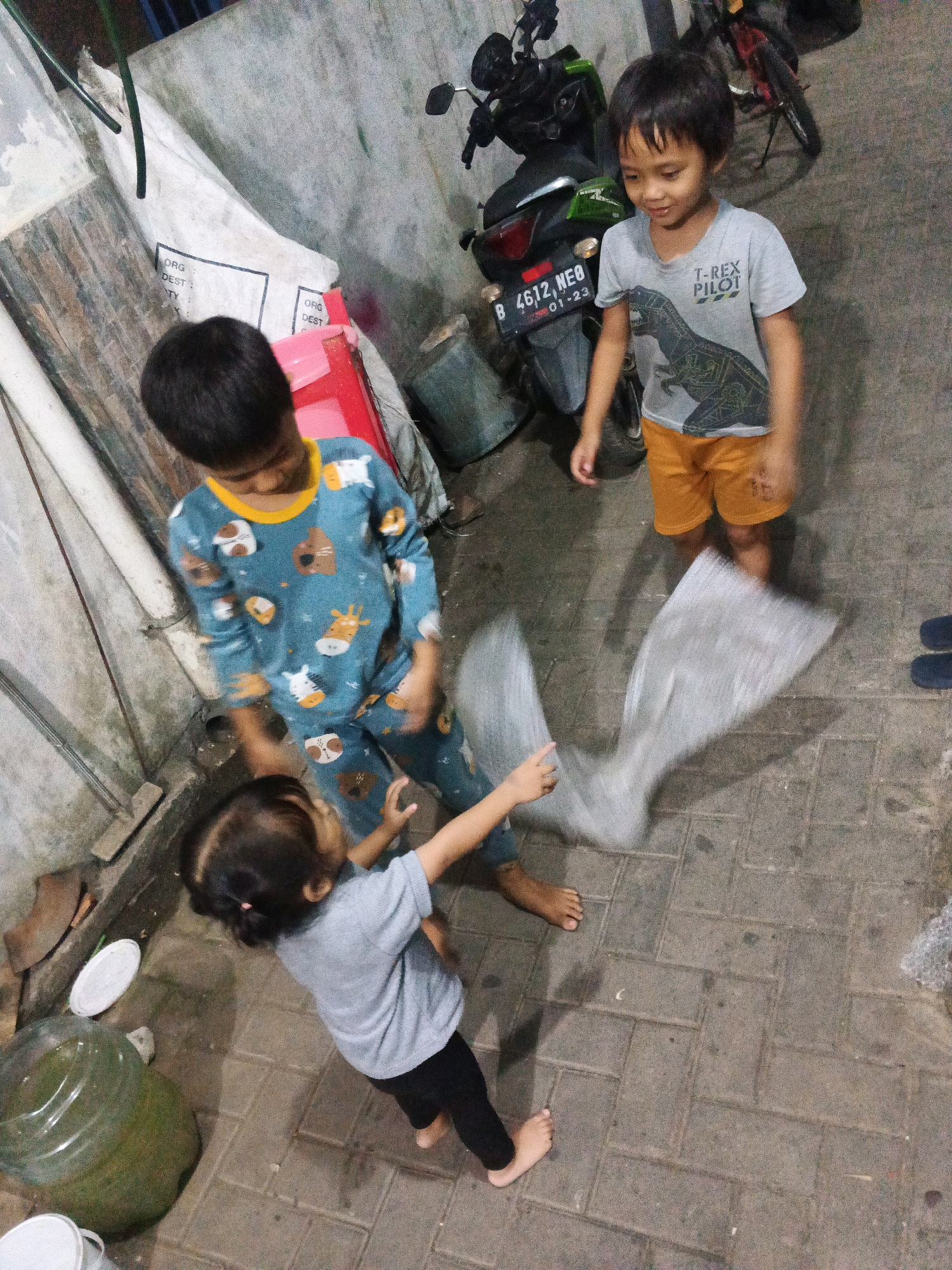 In Kadu, Indonesia, three children are engaged in an activity on a paved ground surface. The child on the left, wearing blue pajamas with animal prints, is standing and looking down at the child in front of them. The child in the center, a young girl in a grey t-shirt and black leggings, is in a low stance, facing the other two children, and holding a piece of transparent, crinkled material. The child on the right, wearing a grey t-shirt with a dinosaur graphic and orange shorts, is standing and looking down at the girl. Behind the children, a motorcycle is parked next to a wall. The environment appears to be an outdoor, possibly residential or communal area, with a rough concrete wall and paved brick flooring. Various objects, including a large plastic water jug and some white containers, are scattered around. The lighting suggests it is either dusk or nighttime.