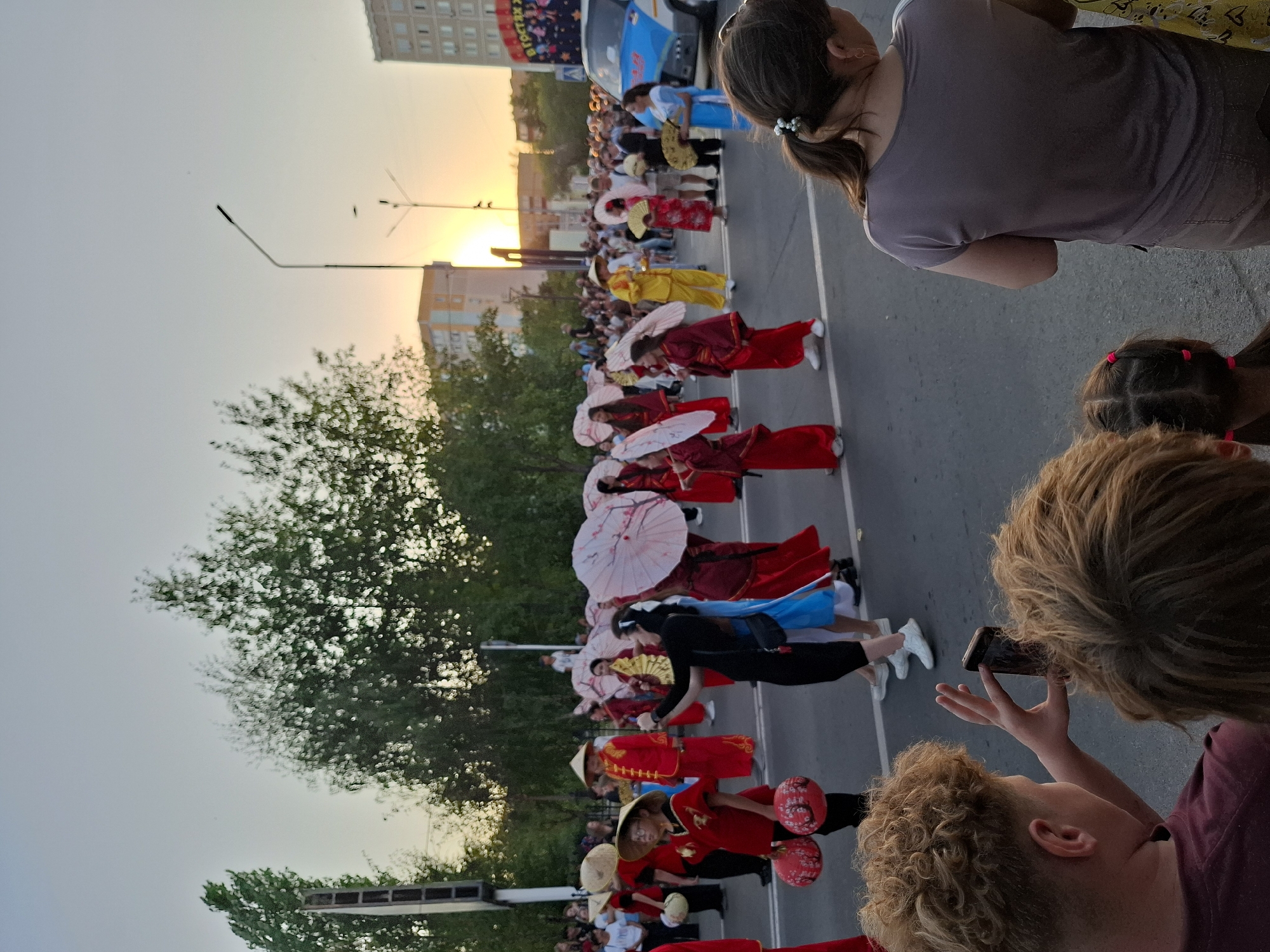 In an outdoor urban setting during sunset, a parade is in progress. The sky is a pale orange and yellow, with scattered trees and buildings visible in the background. The street is paved, with a white line dividing the lanes. A crowd of onlookers lines the street, some standing and others seated. In the foreground, two young people are looking at a smartphone. The middle ground is occupied by parade participants dressed in colorful traditional East Asian attire. Many are wearing conical hats, red robes, or yellow outfits. Some are carrying decorative umbrellas or lanterns. A blue and white van is parked on the right side of the street, and a banner with text is partially visible on its side. The overall atmosphere suggests a cultural festival or celebration.