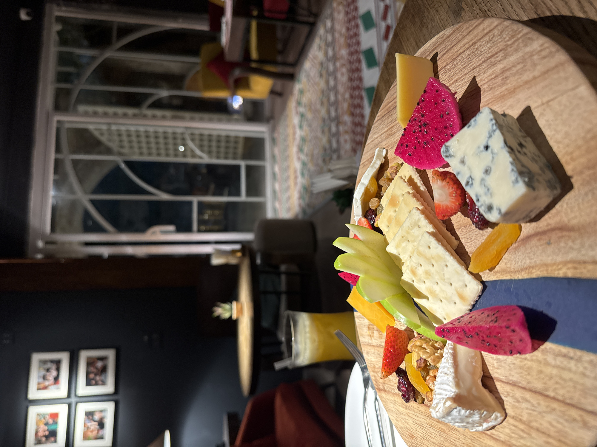 The image shows a close-up of a cheese and fruit platter on a wooden board. The platter includes various cheeses, crackers, strawberries, dragon fruit slices, dried fruit, and nuts. A blue resin inlay runs through the wooden board. In the background, a window with decorative grilles is visible, suggesting an indoor setting with an outdoor view. The overall lighting suggests it might be evening. Further in the background, a patterned rug and some furniture are discernible, hinting at a restaurant or lounge setting. The image appears to be taken in Hanoi, Vietnam, based on the provided location context. No people are clearly visible in the image. The focus is on the food presentation.