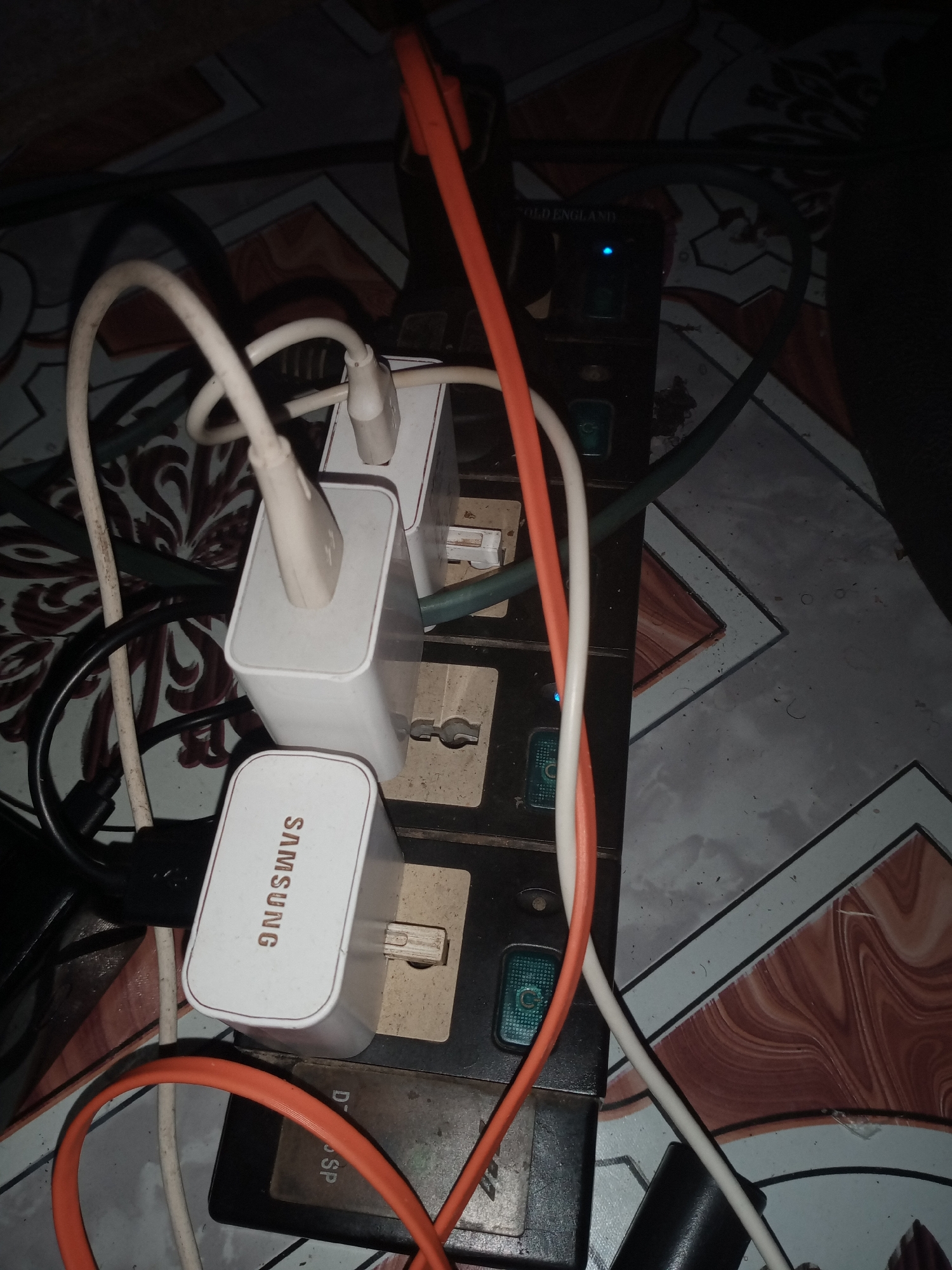 The image shows a close-up view of a power strip plugged into an electrical outlet, located on a tiled floor. Several white and orange cables are connected to the power strip, along with two white charging adapters. One adapter is labeled "SAMSUNG". A small blue LED light is visible on the power strip, indicating it is receiving power. The power strip itself appears to be black and is positioned horizontally. The surrounding environment is dimly lit. The image is taken from a top-down perspective. The location is identified as Jalingo, Nigeria.