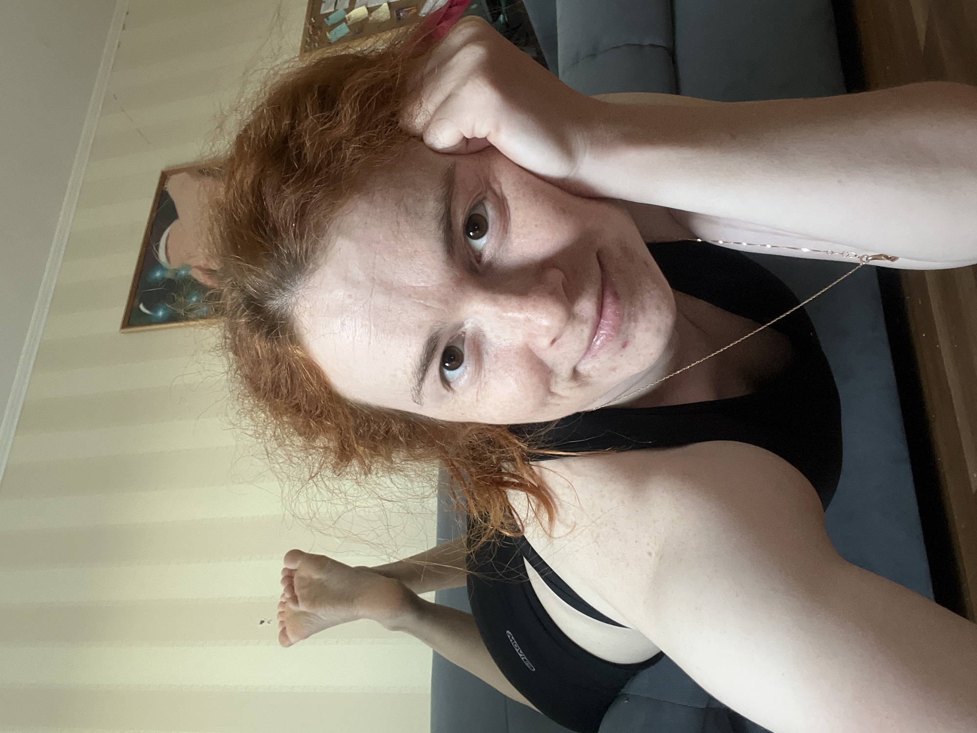 A woman with red, curly hair is lying on a gray couch, looking into the camera. She is wearing a black top. Her right hand is near her forehead. A thin gold necklace is visible. The background shows a striped wall and a framed picture. Part of her left foot is visible. The location in the city of Chaikovsky, Russia, cannot be determined from the image alone.