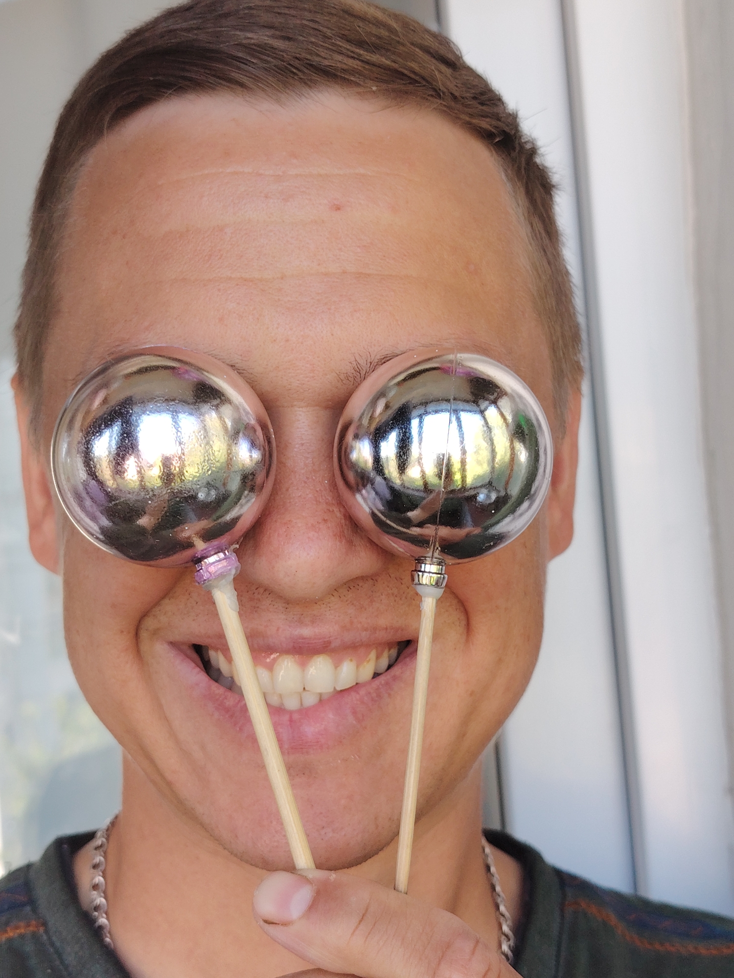 The image is a close-up, portrait-style photograph of a person's face, with two shiny, spherical objects held up to their eyes. The person is smiling, showing their teeth. The background is blurred and appears to be an outdoor setting with a light-colored vertical structure, possibly a wall or door, and some greenery suggesting trees. The lighting indicates it is daytime. The overall impression is playful or humorous. The image was taken in Chaikovsky, Russia.