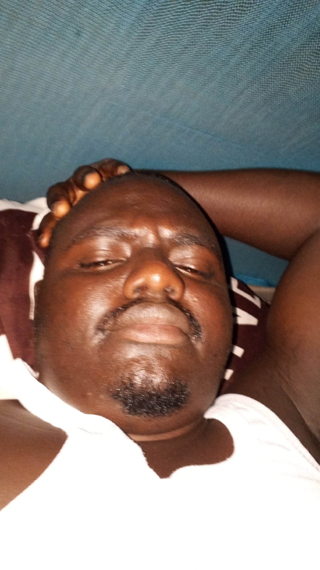 A close-up shot of a dark-skinned man lying down. He has a mustache and goatee, and his eyes are mostly closed. He is wearing a white tank top and appears to be resting his head on his hand. The background consists of bedding and what appears to be a mosquito net. Location: Unknown, Unknown.