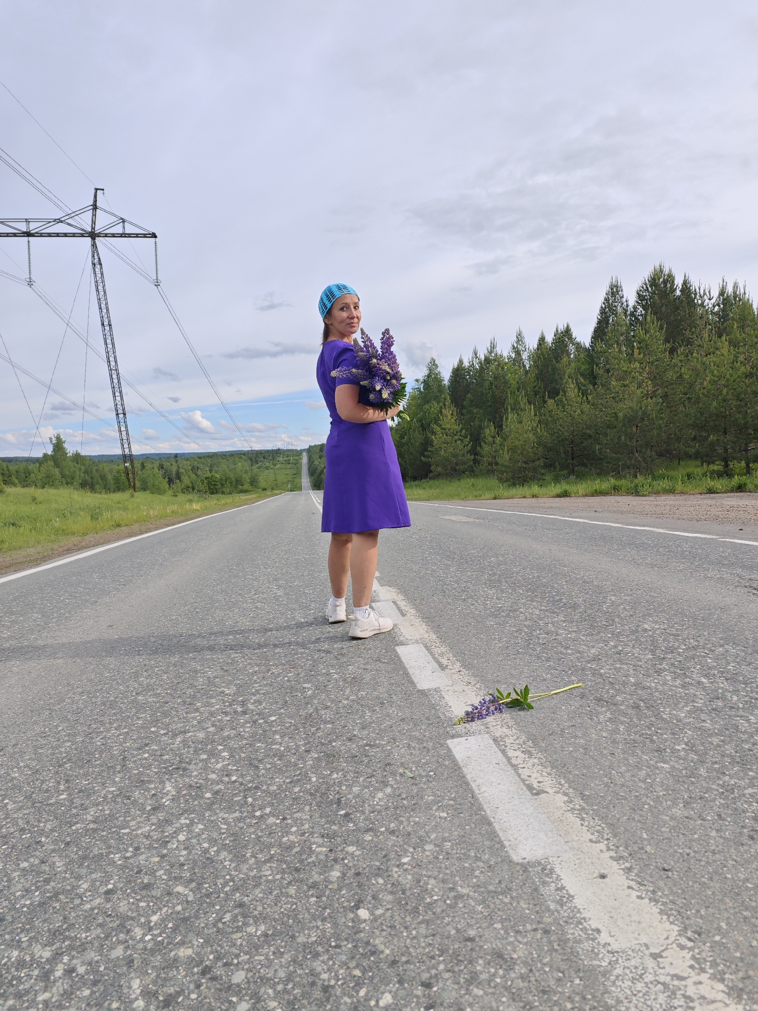 A woman stands in the middle of an asphalt road wearing a purple dress, white sneakers, and a blue crocheted hat. She holds a bouquet of purple lupine flowers. One lupine flower lies on the white line painted on the road. Behind her is a straight road receding into the distance, flanked by green trees and a cloudy sky. A power line structure is visible on the left side of the road. The location is near the city of Chaikovsky, Russia, as suggested by the natural environment and power line infrastructure commonly found in Russian regions.