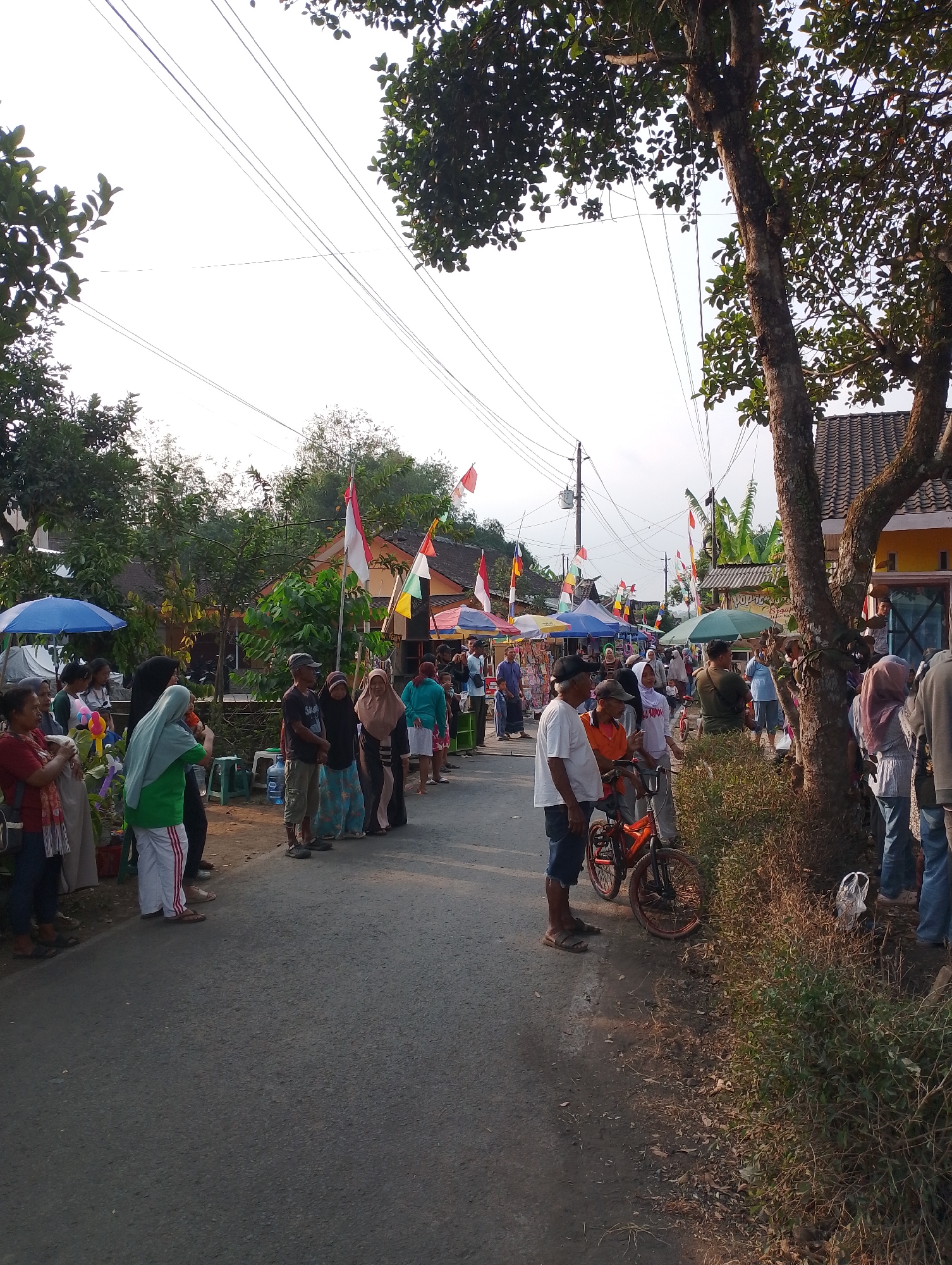 The image depicts a street scene in Krajan Satu, Indonesia, likely during a local event or market. A road runs through the center of the frame, with a line of people gathered on the side. Numerous stalls, some with colorful umbrellas and awnings, are set up along the road, suggesting a marketplace. Indonesian flags are visible, some flying from poles and others attached to structures, indicating a patriotic or celebratory atmosphere.
The people in the image are diverse in age and attire, with many wearing casual clothing. Some individuals appear to be browsing the stalls, while others are standing and conversing. A bicycle is parked by the side of the road, near a group of men. The environment is characterized by trees, houses with tiled roofs, and overhead power lines. The lighting suggests it is either early morning or late afternoon, with the sun casting long shadows. The overall impression is one of community activity and local commerce.