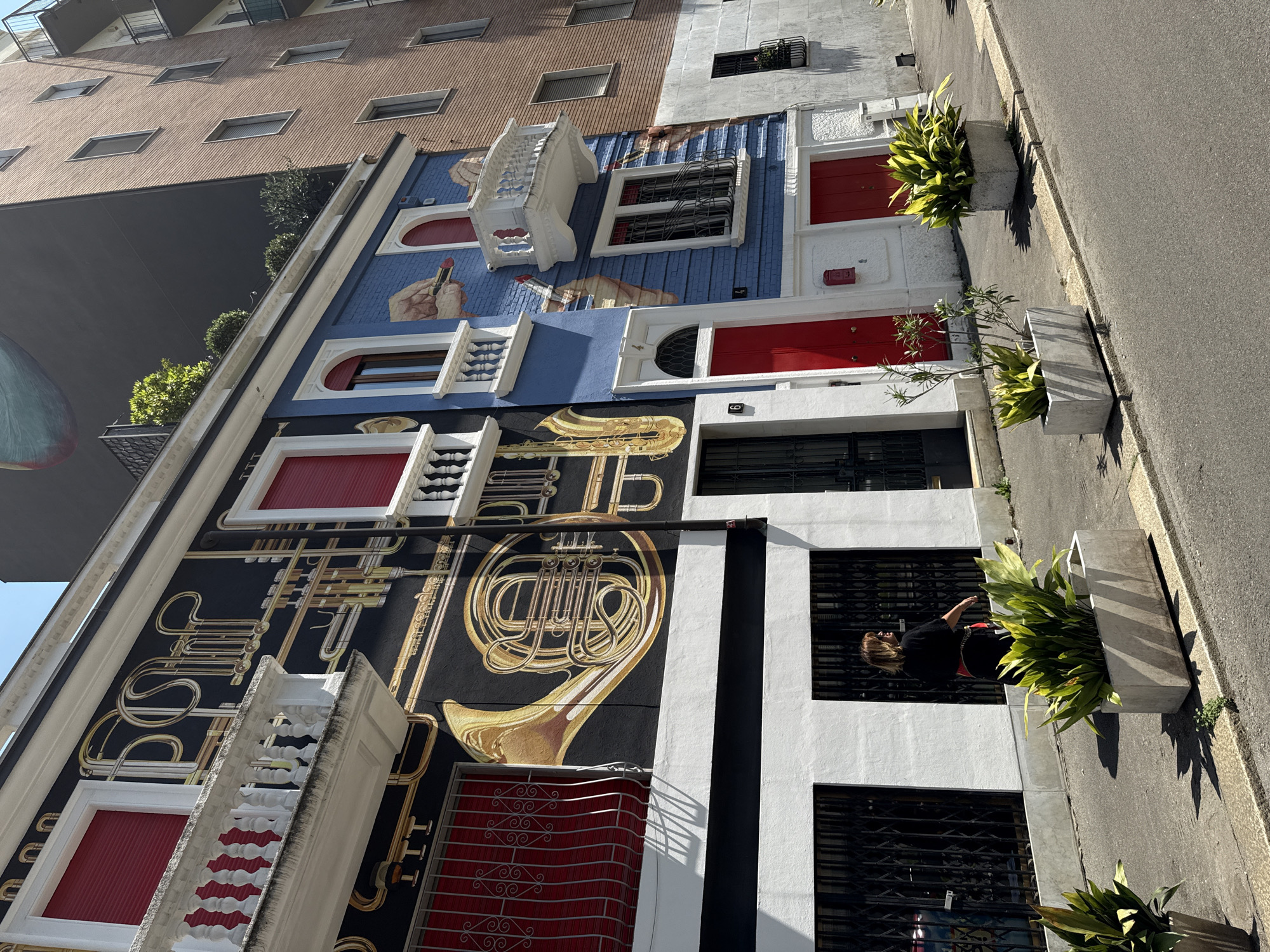 A medium eye-level outdoor shot, shows the "Case delle Arti" (Houses of the Arts) in Milan, Italy, adjacent to a modern brown building. The foreground shows an adjacent street.
The "Case delle Arti" are depicted as a series of adjacent buildings with different facades. From top to bottom, the first facade is painted blue with elements like hands holding a lipstick, balcony elements, red doors and a mail slot. The second facade has a black background with musical instruments depicted on it, with red awnings on the windows and white architectural details. The bottom facade is white with black stripes, black-barred windows and plant containers. A person is standing at the window on the bottom facade, wearing a black top. The street has plant containers along its edge.