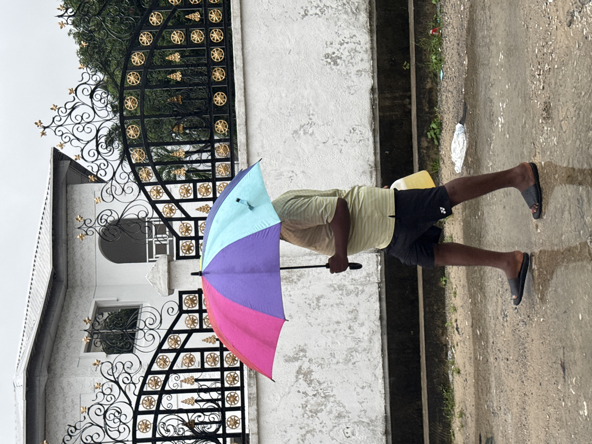 An individual in a striped shirt and shorts is holding a colorful umbrella next to a white building with ornate black and gold gates. The scene appears to be outdoors, with wet pavement visible. The location is Ado, Nigeria.