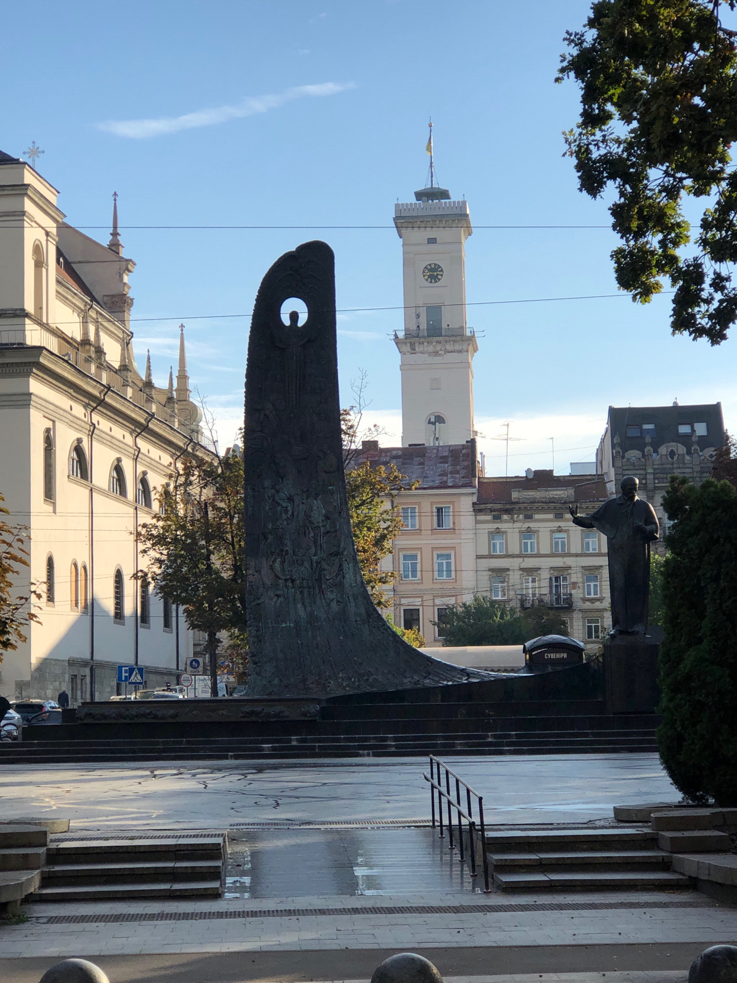 The image captures an outdoor scene in Lviv, Ukraine, featuring a monument and surrounding architecture. In the foreground, a dark, abstract sculpture with a cutout silhouette of a person is prominently displayed. To the right of the sculpture, a bronze statue of a man stands on a pedestal, with his right arm extended. Behind these monuments, a tall, light-colored clock tower rises, its face showing Roman numerals. Adjacent to the clock tower, several buildings with traditional European architecture, featuring arched windows and tiled roofs, are visible. The sky above is blue with a few wispy clouds. The ground in the plaza area appears wet, suggesting recent rain. Trees, including a large one with green leaves on the right side, frame the scene. The overall impression is of a public square or monument site in an urban setting.