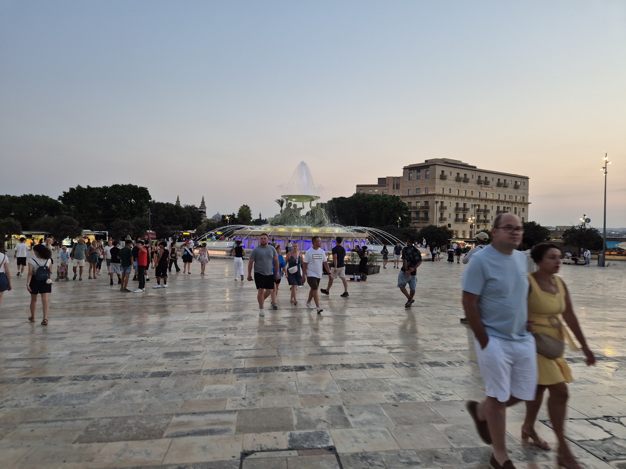 The image depicts a wide-angle view of the Triton Fountain area in Valletta, Malta, during twilight. A large, multi-tiered fountain with sculpted figures and water jets is the central focus, illuminated by lights. Behind the fountain, a multi-story, light-colored building with numerous balconies and windows is visible, likely a hotel or residential building. The foreground is dominated by a vast, paved public square with a stone tile pattern, reflecting the ambient light and showing some wet patches, indicating recent moisture. Numerous people are scattered across the square, walking in various directions. They are dressed in casual summer attire, including shorts, t-shirts, and dresses, suggesting a warm evening. Some are walking individually, while others are in pairs or small groups, engaged in conversations or simply enjoying the public space. In the distance, trees and other architectural elements of the city can be seen, including what appears to be the top of a church dome. The sky is a pale gradient of blue and pink, indicative of the post-sunset period. A street lamp is visible on the right side of the image, further illuminating the area. The scene conveys a lively yet relaxed atmosphere typical of a popular urban gathering spot.