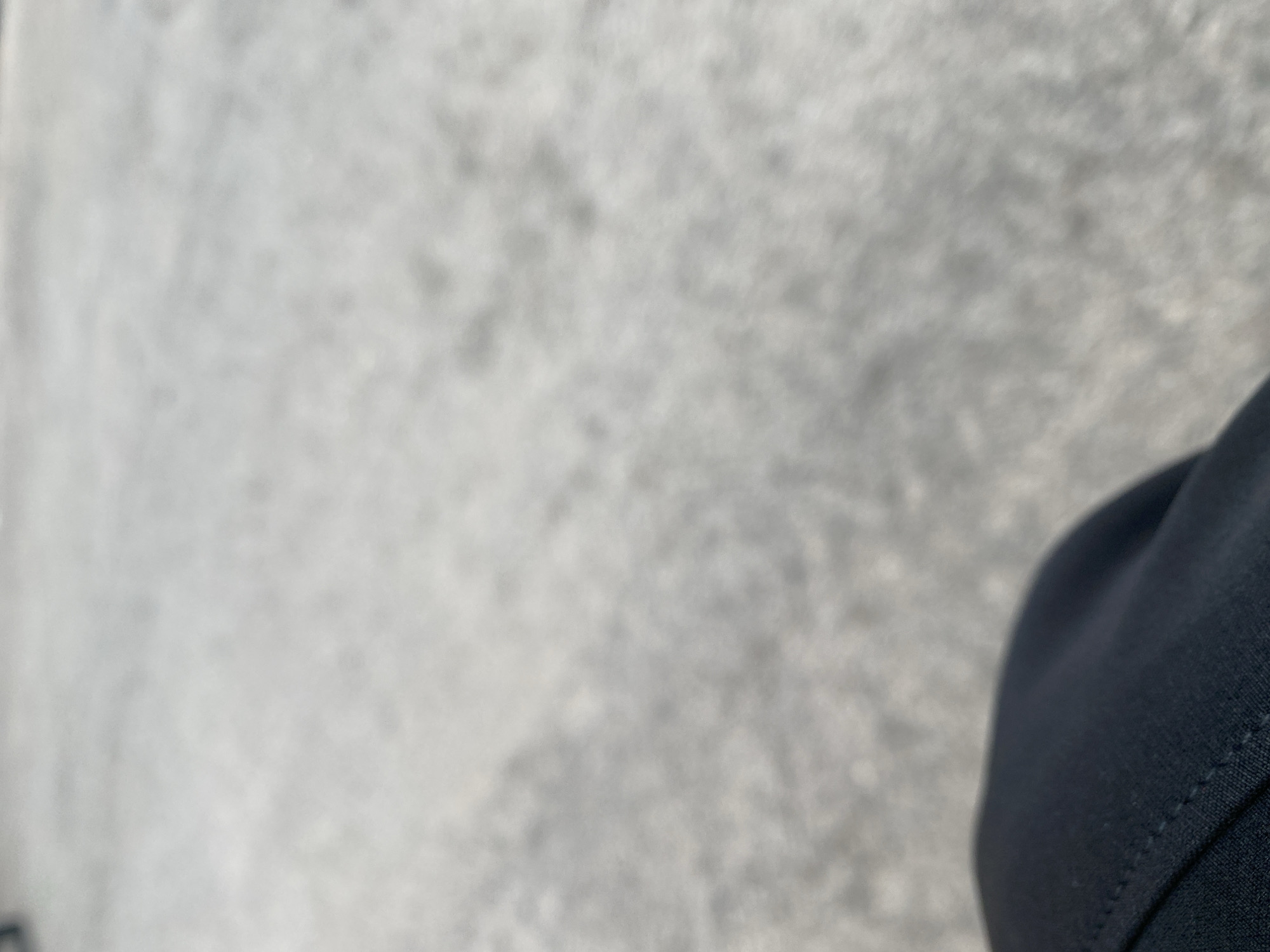 The image shows a close-up view of a dark blue fabric, likely part of clothing, in the lower right corner. The rest of the frame is filled with a blurred, speckled grey surface, possibly pavement or a textured wall. There are no discernible people, specific objects, or clear scene details due to the extreme blur. The lighting appears to be diffuse, suggesting an overcast day or shade. No text is visible. The context of Jalingo, Nigeria, provides a geographical location but no specific visual cues within the image itself can confirm this. The overall impression is abstract due to the lack of focus and clear subject matter.