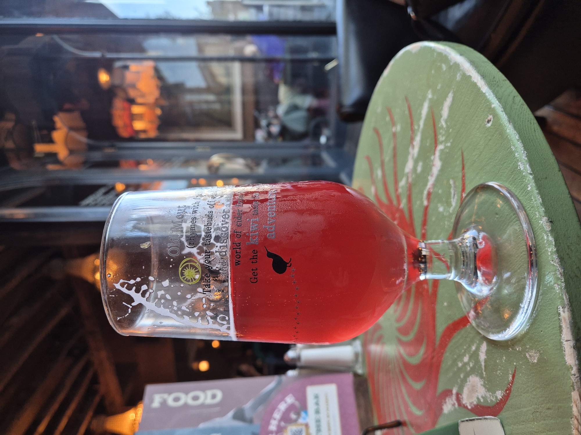 A sideways close-up shows a partly-filled glass of red liquid sitting on a circular green table top with a red sunburst design. The glass has writing on it and what appears to be a kiwi bird logo. The glass is labeled "Old Mout" with the tagline "Get the Kiwi adventure". In the background, blurred out are windows showing a city street scene. The scene appears to be inside a pub or restaurant in West Suffolk, United Kingdom. A portion of a menu is visible at the bottom.