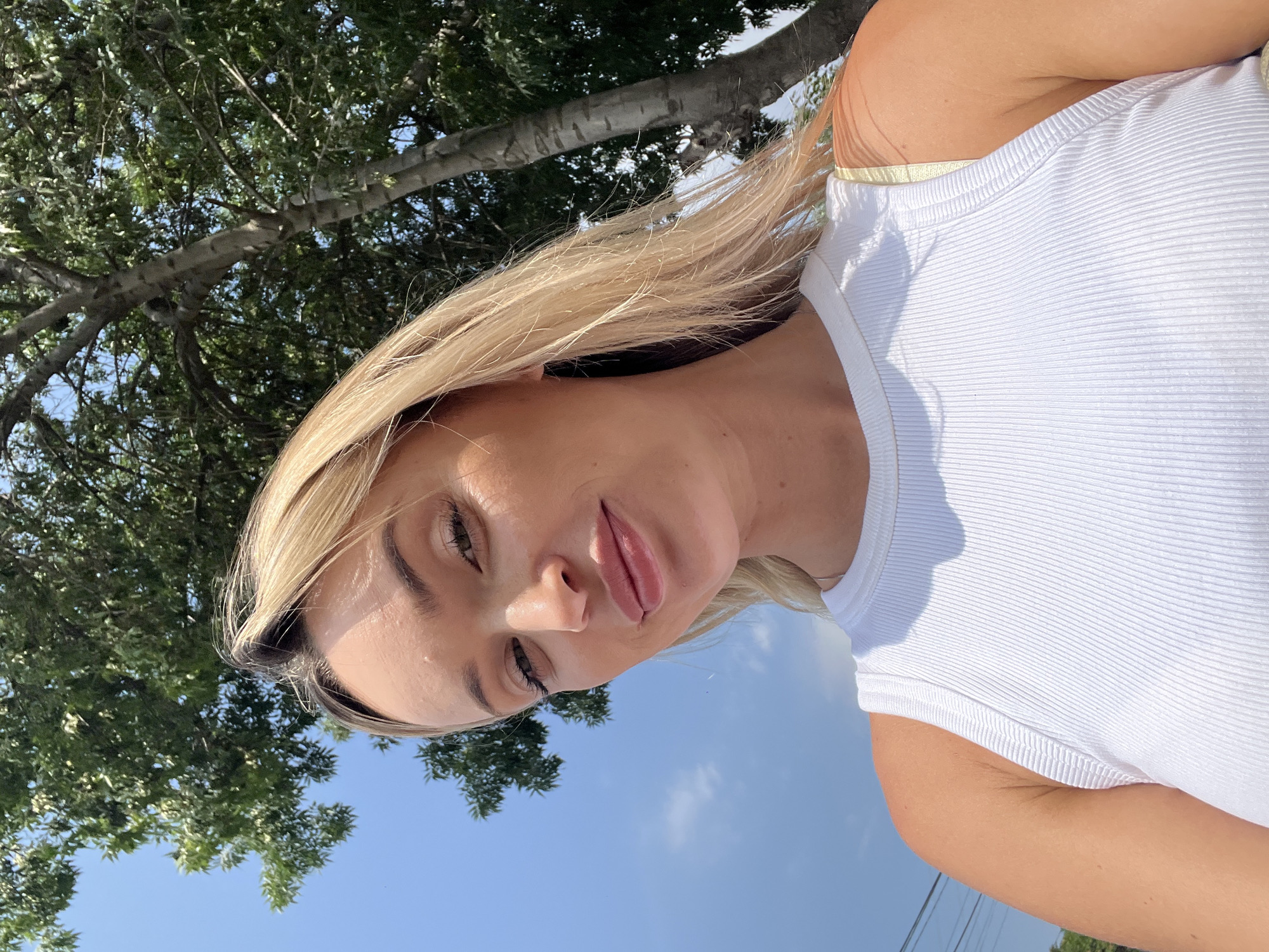 A young Caucasian woman with blonde hair is shown from the chest up, looking into the camera. She is outdoors, with a bright blue sky and green trees visible behind her. The woman is wearing a white ribbed tank top. The sun is shining brightly, casting shadows on her face and the trees. She has on light makeup, including mascara and lip gloss. The setting appears to be a sunny day in a park or wooded area, possibly in the city of Fontanka, Ukraine, though this cannot be definitively confirmed from the image alone.