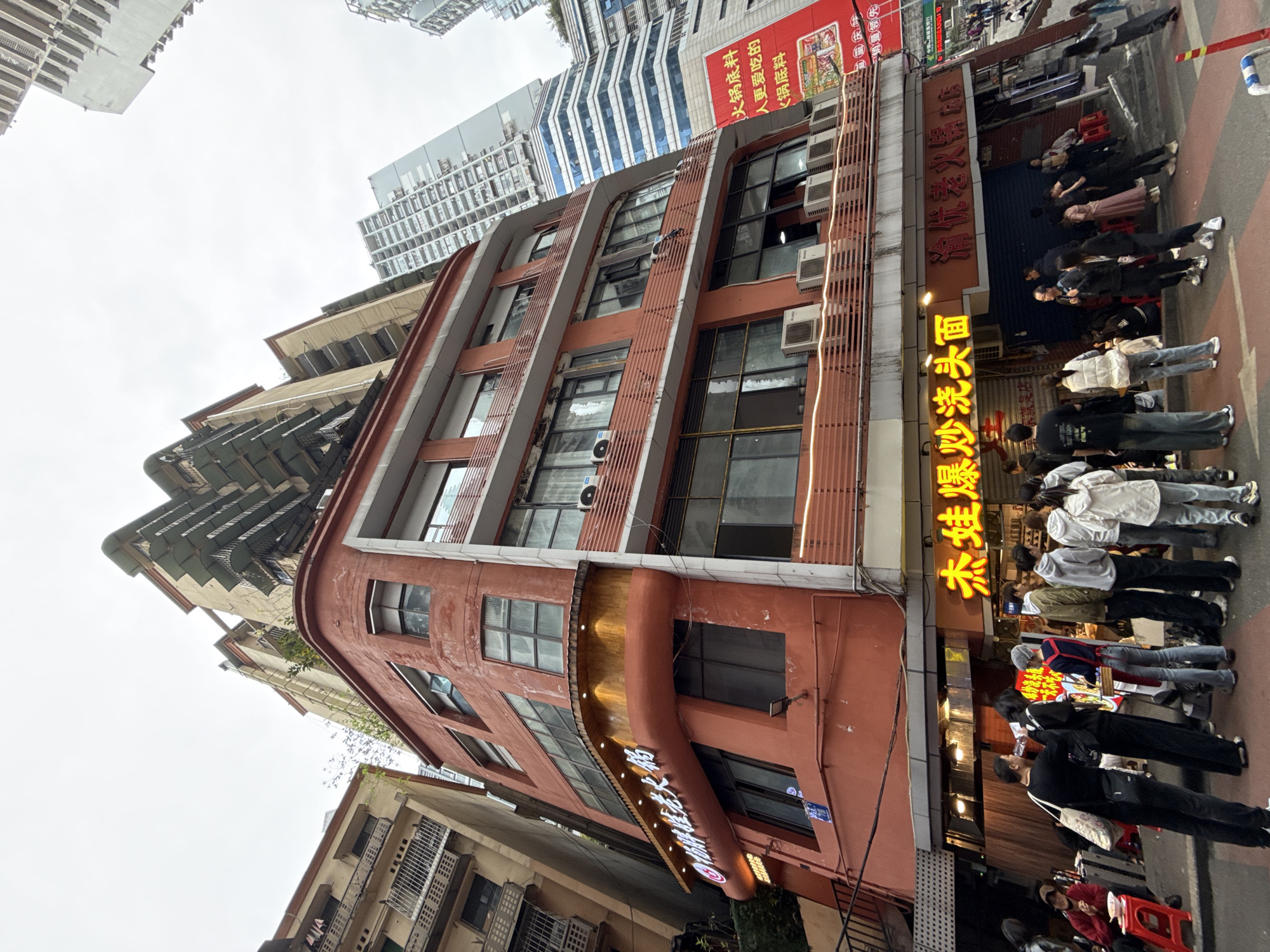 The image presents an urban street scene in Yuzhong District, China, under an overcast sky. A prominent five-story building, painted in a rust-orange hue with a rounded corner, dominates the foreground. Its facade features numerous windows, some ajar, and multiple exterior-mounted air conditioning units. The ground floor accommodates two eateries: one identified by a bright yellow neon sign reading "杰娃爆炒浇头面" (Jiewa Baochao Jiaotou Mian), and another with a red sign displaying "渝怀老火锅店" (Yuhuai Laohouguo Dian). Wooden paneling with recessed lighting extends above the main storefronts.
Approximately 20 to 30 individuals, primarily young adults in casual attire, are present on the sidewalk in front of these establishments. A queue has formed at the entrance of "Jiewa Baochao Jiaotou Mian," indicating active business. Several patrons are seated on small red plastic stools at low tables directly on the sidewalk, consuming food. One individual, possibly an employee, stands near the entrance holding a tablet or menu. To the immediate left, an older, multi-story building with metal balconies ascends a slope. In the background, a cluster of modern high-rise buildings, some with blue-tinted glass facades, contrasts with the older architecture. A large red billboard advertising hotpot is visible on the right, further down the street. The street surface exhibits pedestrian markings and a low blue-and-white barrier.