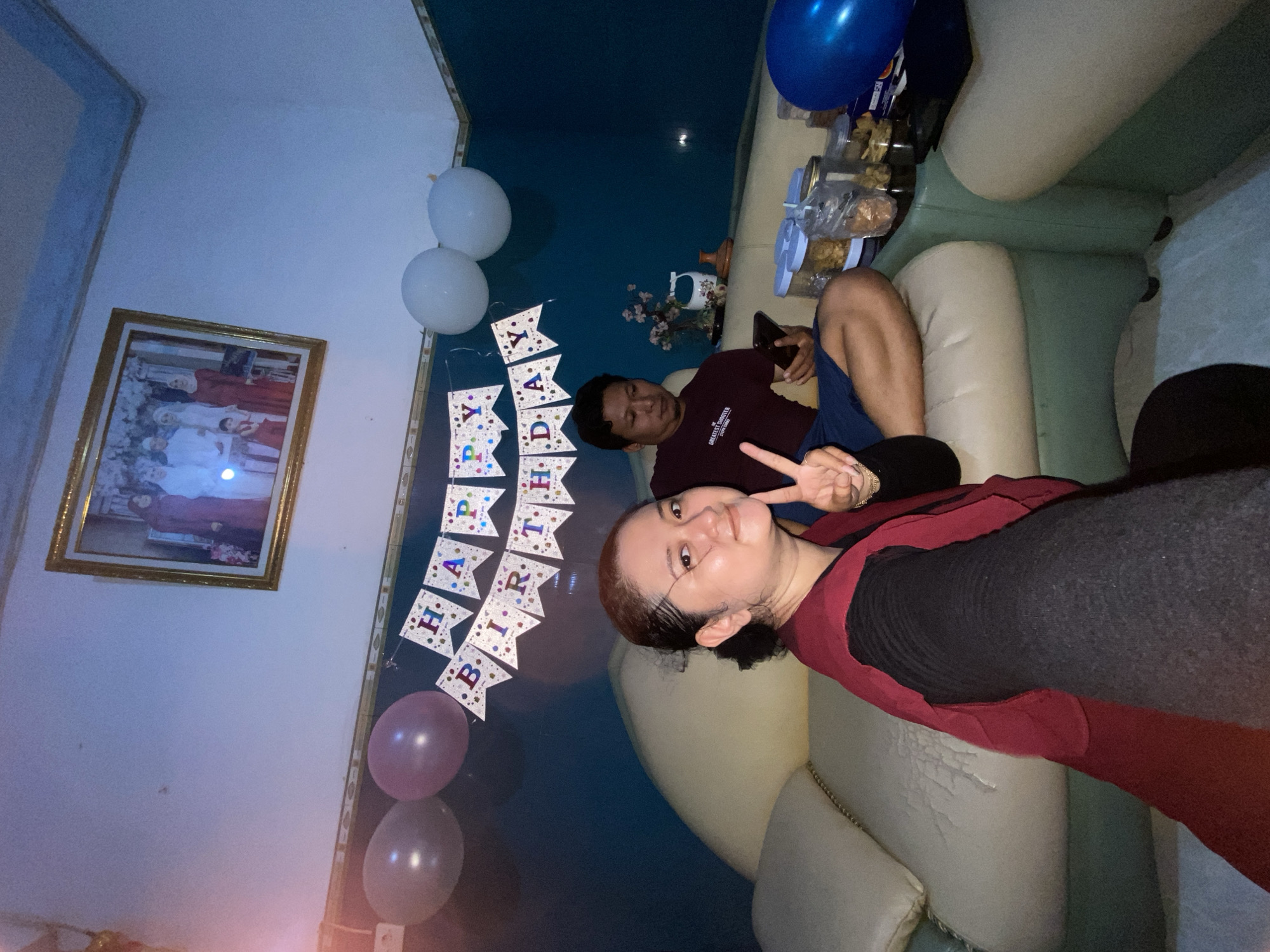 Gelam, Indonesia. A woman poses for a selfie while giving a peace sign. A man is visible on the sofa. A “Happy Birthday” banner is in the background, along with balloons. A picture frame is on the wall. Jars of food are on the sofa near a blue balloon. The sofa is beige.