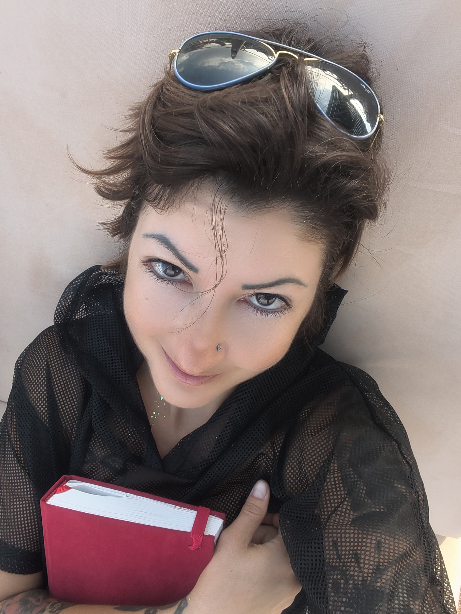 A woman with dark, tousled hair, wearing aviator sunglasses perched on her head, lies on a light brown surface. She is wearing a black mesh top and holding a red notebook. Her head is tilted slightly, and she is looking up at the camera with her eyes open. There are specks of green glitter on her chest and a nose ring. The background appears to be a muted, textured surface. This image was taken in Nahariyya, Israel.
