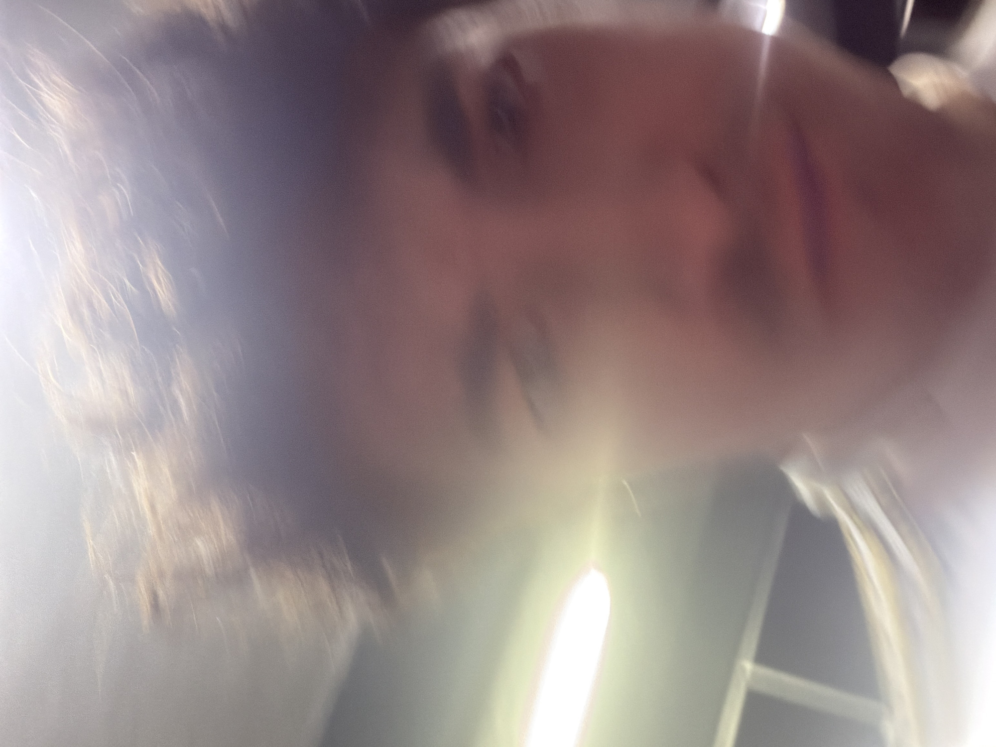 The image is a first-person perspective, selfie-style photograph with a shallow depth of field, causing much of the image to be out of focus. The subject appears to be a young person, likely male, with brown hair. Their face is the primary focus, though it is blurred. The lighting is bright, suggesting an indoor environment with artificial light sources. The background is indistinct but hints at a modern interior with some structural elements visible. There are no clear indications of the city of Cancún, Mexico, or any specific landmarks. The overall impression is that of a casual, candid shot taken with a mobile device.