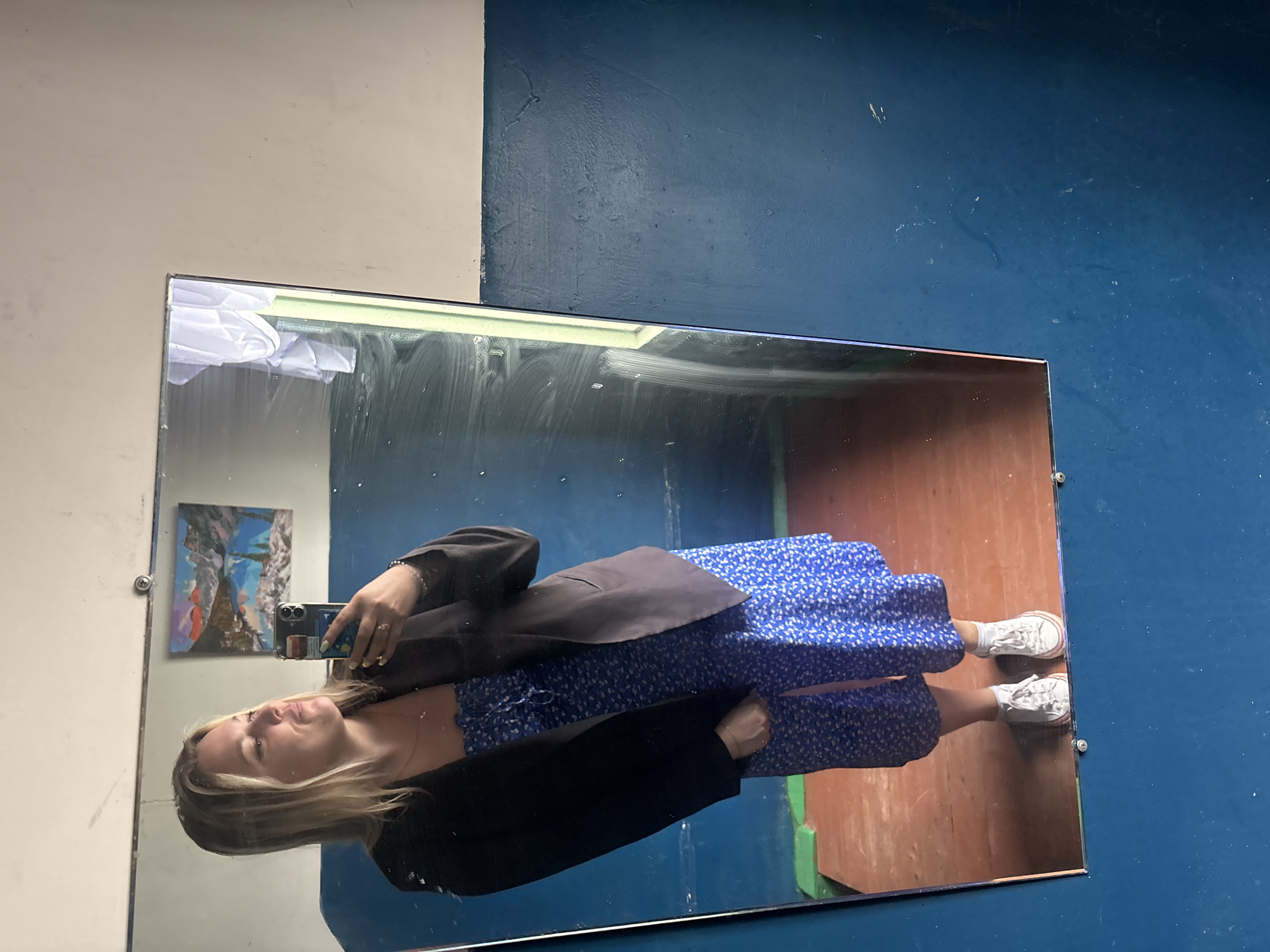 A woman stands in front of a mirror, taking a selfie. She is wearing a black blazer over a blue floral-print dress and white sneakers. In the reflection, a painting of a mountain landscape is visible on the wall behind her. The room appears to be a simple, possibly unfinished space, with blue-painted walls and a wooden floor. The mirror itself has some smudges and streaks on its surface. The overall setting suggests a casual, personal moment captured in what might be a residential or backstage area. There is no specific information to place this scene in Chaikovsky, Russia.