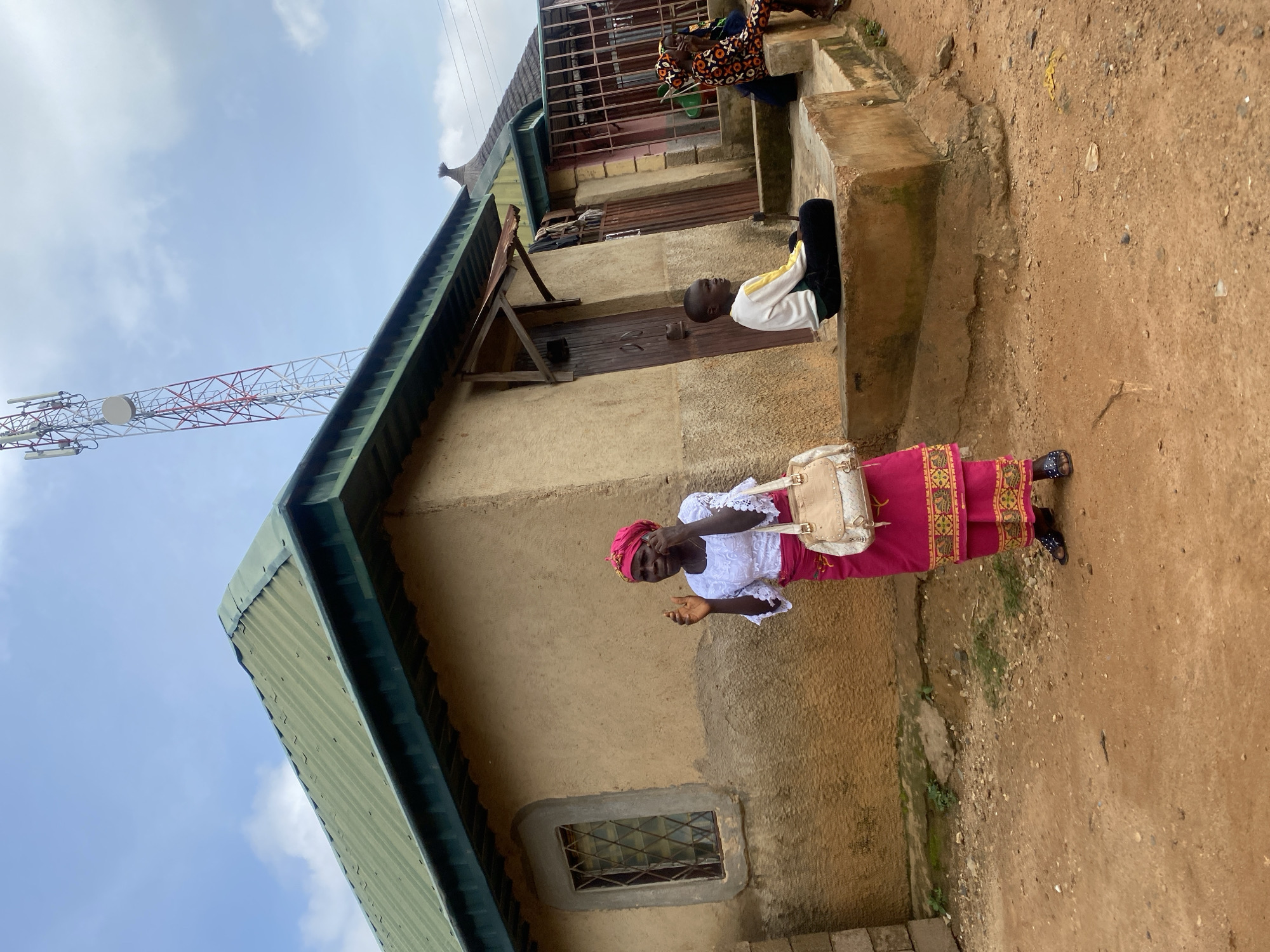 A woman in a pink headscarf and a white lace top with a matching pink skirt is standing on a dirt path outside a building. She is holding a beige purse on her left shoulder and is speaking on her mobile phone, with her right hand raised. The building appears to be made of mud or stucco with a corrugated metal roof. To the right of the woman, a young boy in a white and yellow long-sleeved shirt and black pants is sitting on a concrete step. Further to the right, a person in a colorful patterned outfit is sitting on the steps, and another person is partially visible behind them. A tall telecommunications tower is visible in the sky behind the building. The overall environment appears to be rural or a peri-urban area in Rafin Par, Nigeria, with a dusty ground and simple structures.