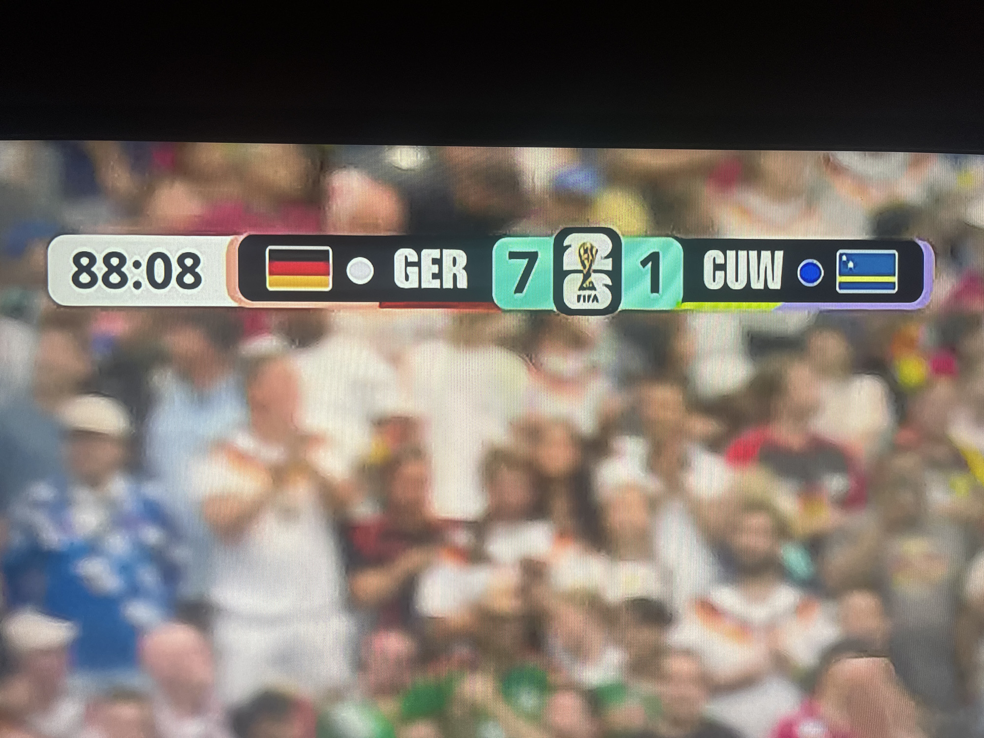 This is a close-up, angled shot of a scoreboard at a sporting event, likely football, displayed on a large screen or monitor. The scoreboard shows the time as 88:08. On the left side of the scoreboard, the German flag is displayed, followed by the abbreviation "GER" and the score "7". In the center, between the two scores, is the FIFA World Cup trophy logo. To the right of the trophy is the abbreviation "CUW" and the score "1", followed by a circle and the flag of Curaçao. The scoreboard is superimposed over a blurred background of spectators in a stadium. The audience members are diverse in age and are wearing casual clothing, with some appearing to be wearing team colors. The overall scene suggests a live sporting match, possibly a World Cup qualifier or a related event, given the presence of national flags and the FIFA trophy. There is no indication that this scene is located in the city of Ruse, Bulgaria.