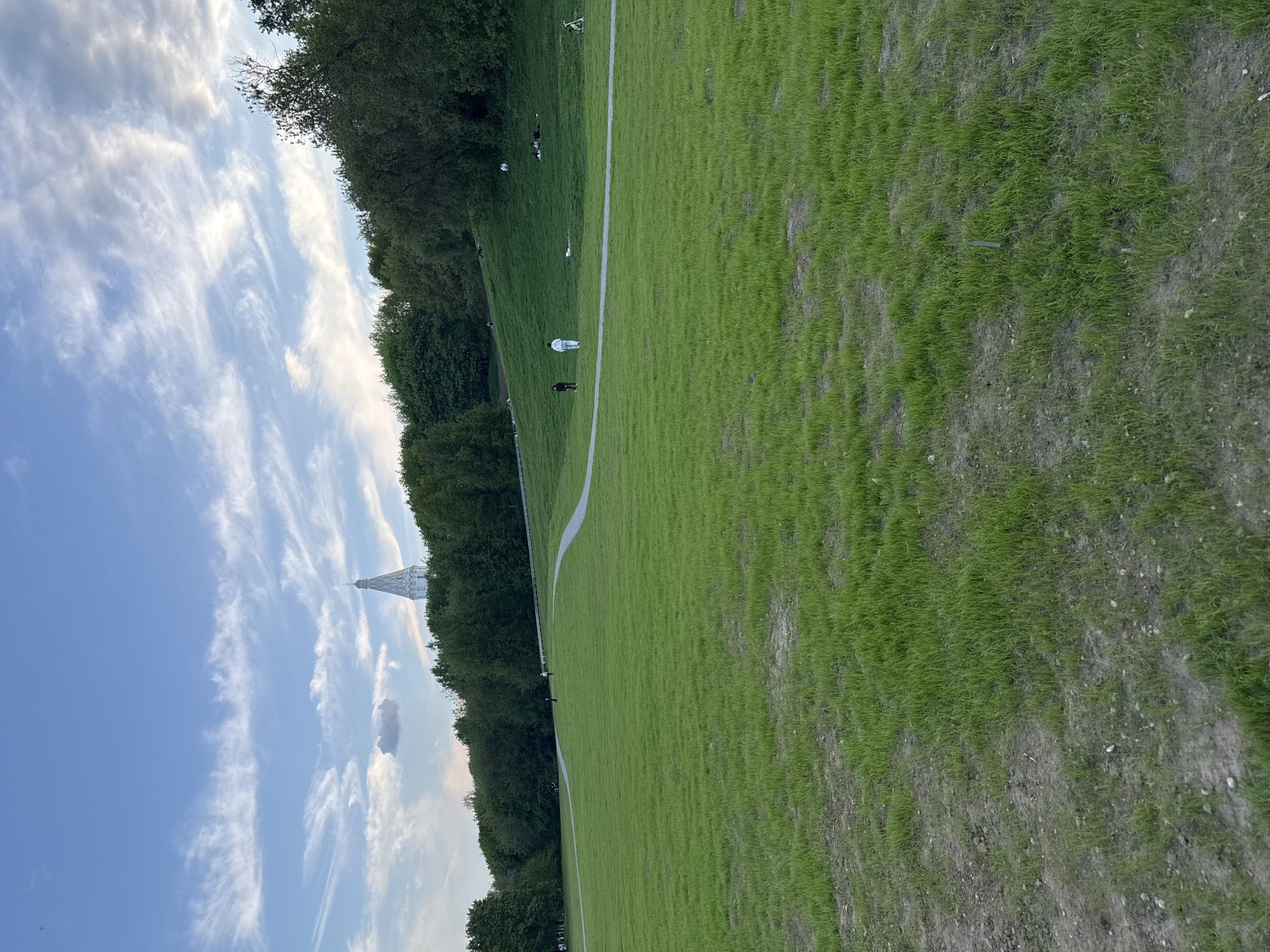 The image shows a grassy field in Moscow, Russia, with the Church of the Ascension at Kolomenskoye visible in the distance. A path winds through the field, and several people can be seen walking along it. The sky is blue with scattered clouds. The grass is green and lush in some areas, while other areas show patches of dry soil.