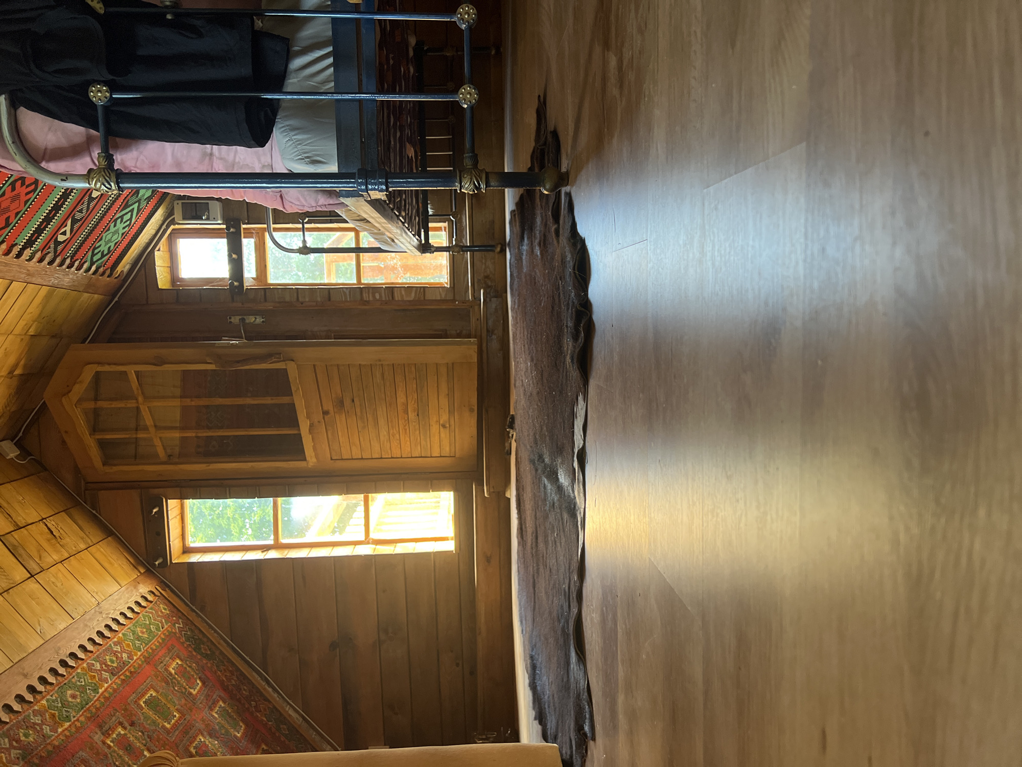 The image depicts the interior of a room with wooden walls and a slanted wooden ceiling, likely an attic space. The floor is light-colored wood laminate. A dark, shaggy rug is placed on the floor in the mid-ground. To the right, there is a dark metal bed frame with a pink duvet and black clothing draped over it. Two tall, narrow windows are visible on the back wall, letting in natural light. A wooden door with glass panes is positioned between the windows. A colorful patterned rug is draped on the slanted ceiling on the left side of the room, and a similar rug is partially visible on the back wall behind the bed. The scene appears to be a cozy, rustic living space. This room is located in the city of Norviliškės, Lithuania, as part of the Norviliškės Manor complex.