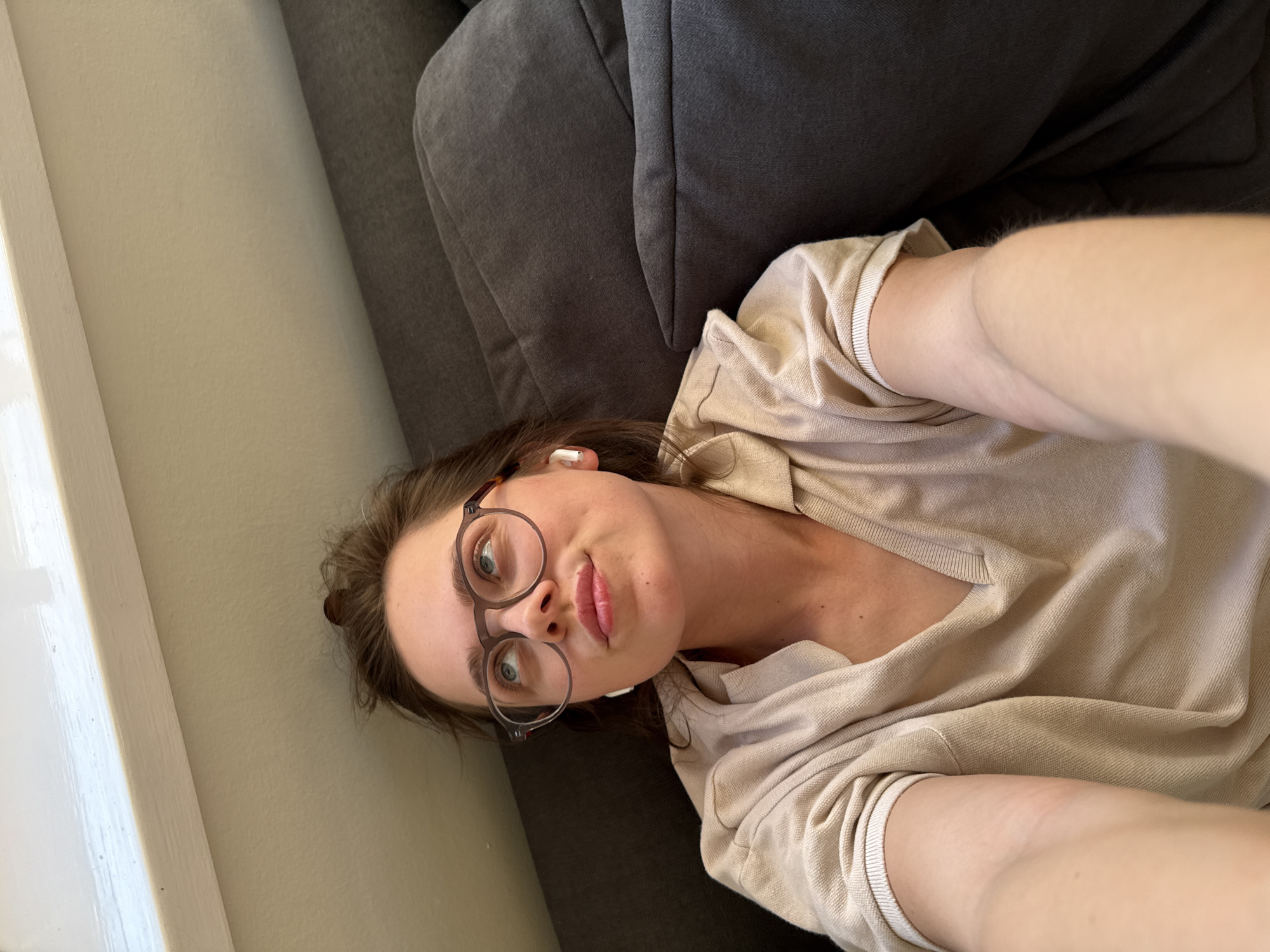 A selfie captures a young woman lying down on a dark grey sofa indoors, in what appears to be a casual setting in Budapest, Hungary. The woman, likely in her 20s or 30s, has fair skin and visible long brown hair. She is wearing round, tortoise-shell framed glasses that accentuate her light-colored eyes. A white wireless earbud is visible in her right ear. Her expression is neutral with slightly pursed lips as she looks directly at the camera. She is dressed in a light beige or tan collared, long-sleeved shirt, possibly a knit polo.
The photo is taken from a low angle, with the woman's head tilted towards the left side of the frame, indicating she is lying on her back or side. To her left is a plain light beige wall with a prominent white window frame or trim, suggesting natural daylight is illuminating the scene. The dark grey fabric of the sofa provides a muted background. The overall atmosphere is relaxed and informal, set during daytime. No text is visible in the image.