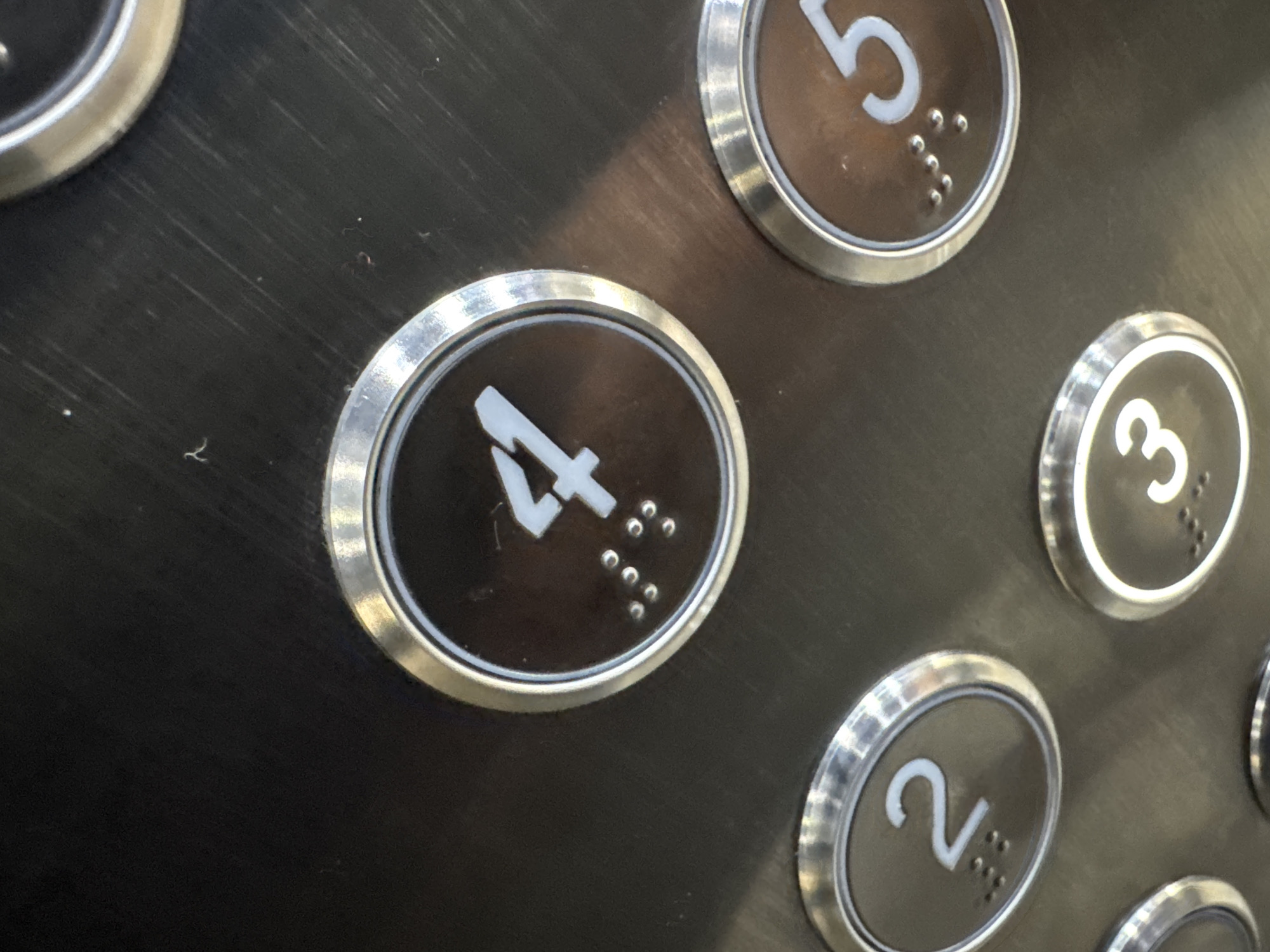 The image shows a close-up view of an elevator control panel, likely in Koltsovo, Russia. The panel is made of brushed metal and features several circular buttons with illuminated numbers and braille. The visible buttons include "4," "2," and "5," and a partial view of a button labeled "3" is also present. Each button is surrounded by a silver-colored trim. The button for the number "4" is centrally located and is the most prominent. It displays a large white numeral "4" and a series of braille dots below it. To the left of the "4" button is a button for "2," and to the right is a button for "5." The "3" button is visible towards the bottom of the frame. The overall environment appears to be indoors, within an elevator car. There are no people visible in the image. The metal surface of the panel shows some minor scuff marks and dust.