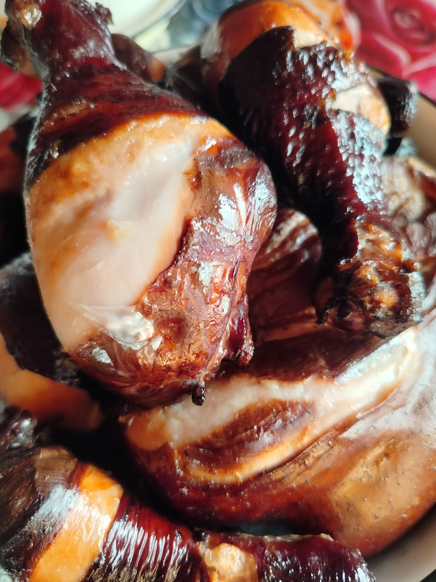 The image shows a close-up, overhead view of several cooked chicken drumsticks piled in a dish. The chicken has a dark, glossy skin, indicative of being smoked or glazed. Some of the meat is visible where the skin is pulled back, appearing pale and moist. The dish itself is not fully visible, but a hint of a floral pattern, possibly a tablecloth or the rim of the dish, can be seen in the upper left and right corners. There are no people visible in the image. The lighting appears to be natural or indoor lighting, creating highlights and shadows on the chicken. The scene is static and focused solely on the food. The image does not provide any information about its location in Kurlek, Russia.