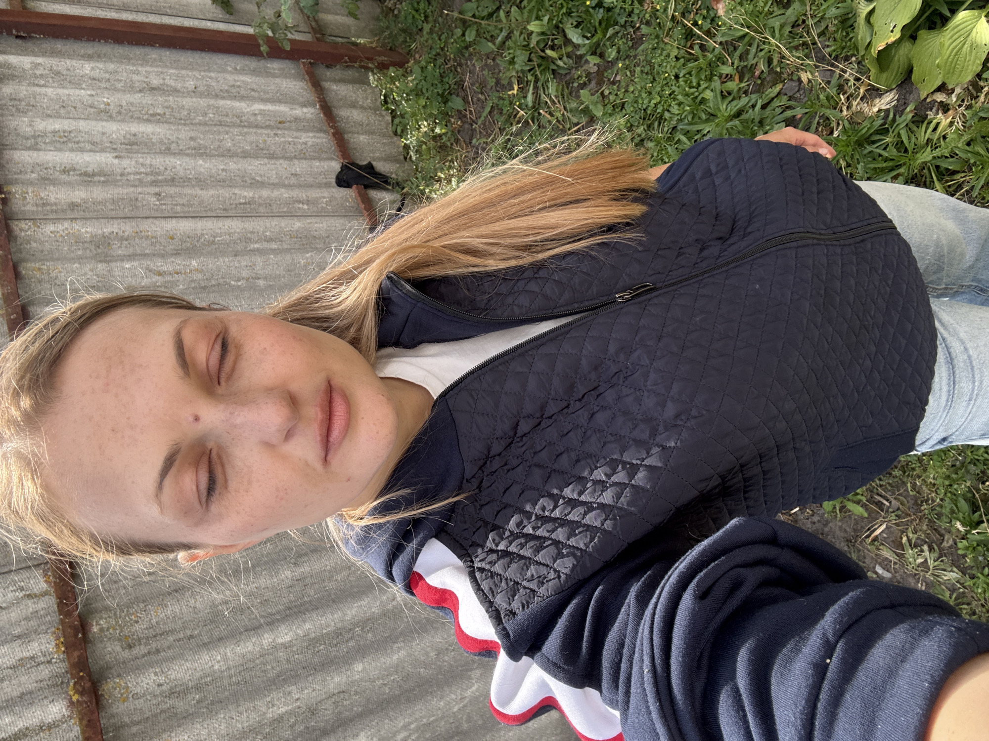 A blonde woman is taking a selfie outdoors. She is wearing a dark blue quilted vest over a white t-shirt and jeans. The vest has red and white trim on the shoulders. Her eyes are closed and she has freckles on her face. Behind her is a grey corrugated fence and some green foliage. The scene appears to be in a rural or suburban area, possibly in Fominka, Russia, given the fence and vegetation. The lighting suggests it is daytime.