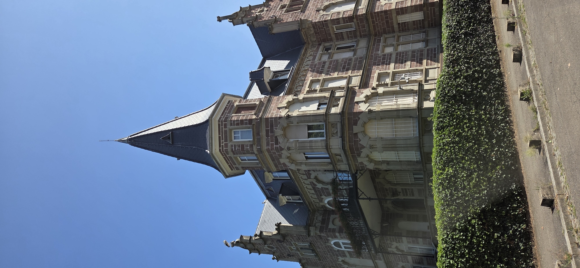 The image shows a large, ornate building with a steeply pitched dark roof and a prominent turret. The building is constructed with red stone and has many windows, some with arched tops. A balcony with an elaborate metal railing extends from the second story. A dense green hedge runs along the front of the property, obscuring the lower part of the building. The sky is clear and blue. In the foreground, a paved path or road is visible with a concrete curb and several small concrete blocks placed at intervals. A white stork is perched on the roofline to the left of the turret. This building is located in Strasbourg, France.