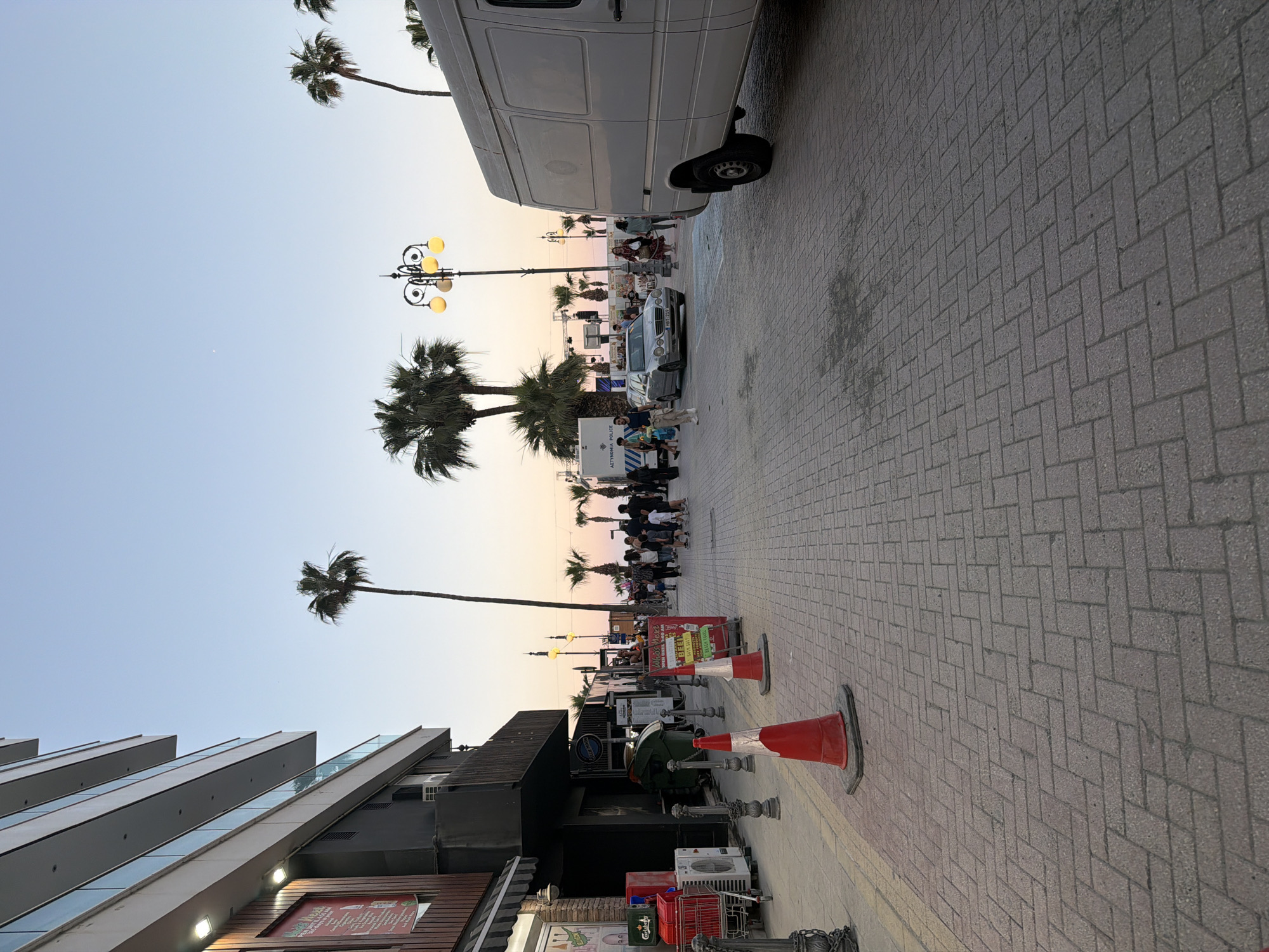 The image shows a street scene in Larnaca, Cyprus. The street is paved with herringbone-patterned grey bricks. A white van is parked on the right side of the street. On the left, there is a multi-story building with a modern design. Palm trees line the street. A group of people is gathered in the distance. Orange and white traffic cones are visible on the sidewalk. Streetlights with globe-shaped lamps are placed along the street. The sky is light blue and orange, suggesting dusk or dawn.