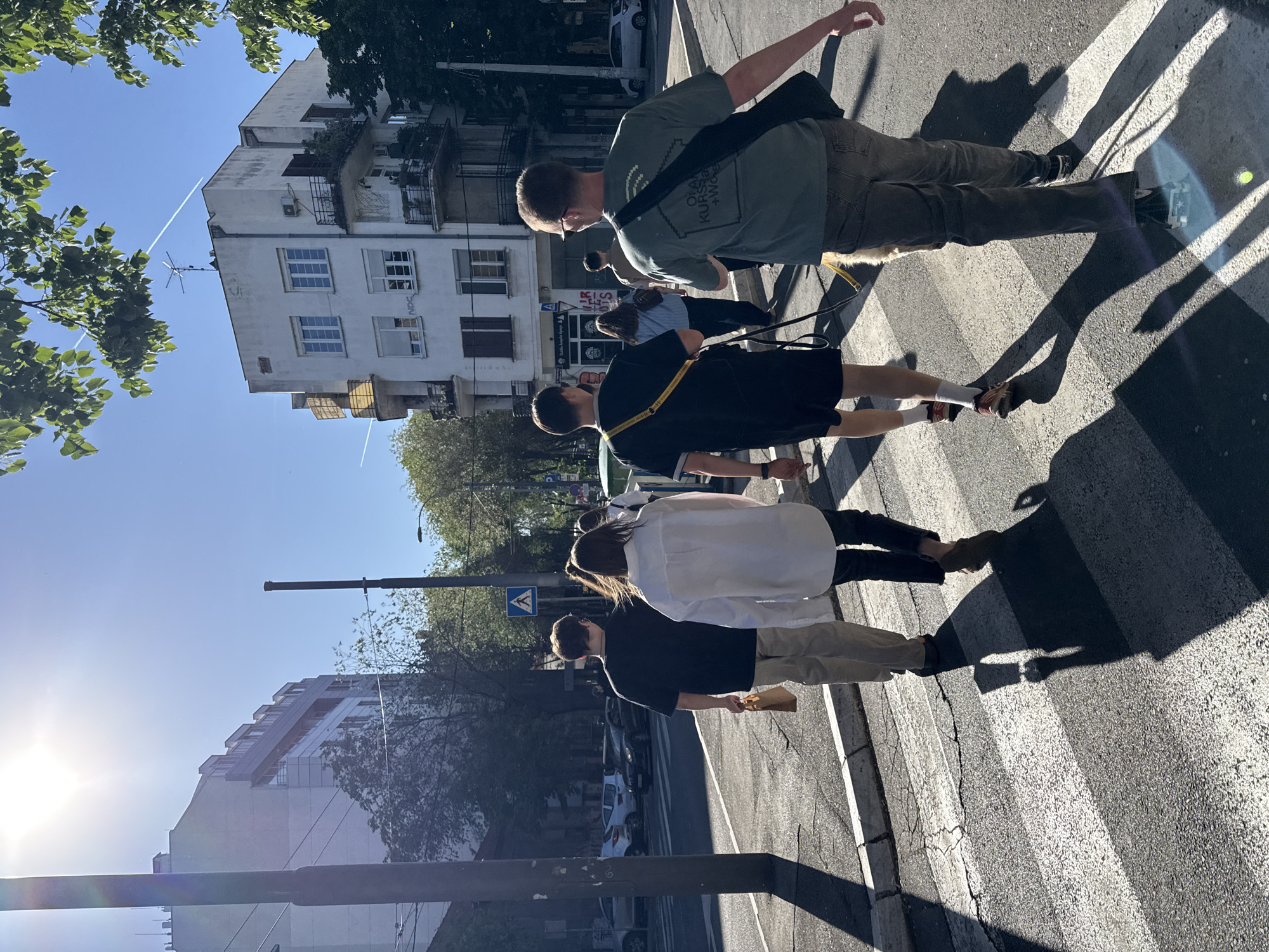 A group of people are crossing a zebra crossing in Belgrade, Serbia. The location features buildings on both sides of the street, trees, a traffic sign, and parked cars. The sky is blue and clear. The group consists of at least four individuals. The first person visible is wearing a green t-shirt and khaki pants. The second person is wearing a black dress with a yellow strap. The third person is wearing a white shirt and khaki pants. The fourth person is wearing a black t-shirt and khaki pants. The street and the buildings show signs of urban wear and tear. The sun casts long shadows from the people and objects on the street.