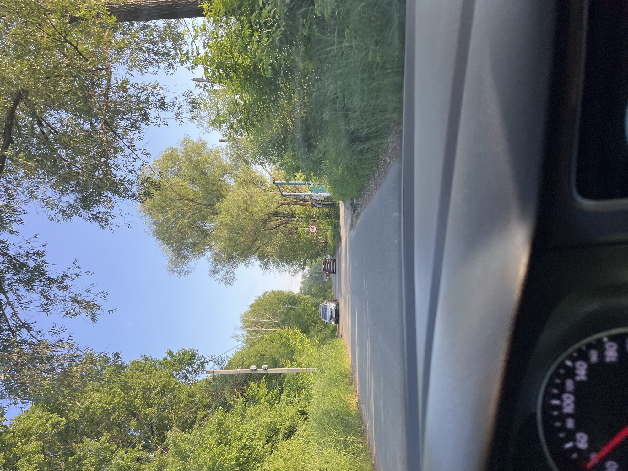 The image is taken from the interior of a car looking forward. A paved road with a single lane in each direction curves to the left ahead. Dense green trees and vegetation line both sides of the road. A power pole with electrical equipment is visible on the left side of the road. Several cars are on the road ahead; a silver SUV is closest in the left lane, followed by a dark-colored sedan in the right lane. A circular speed limit sign with "40" is posted on the right side of the road. The sky is clear and blue. The dashboard of the car is visible in the foreground, with a speedometer showing a speed below 60 km/h. The scene appears to be on the outskirts of a city or in a rural area, given the extensive greenery and the absence of urban infrastructure. Based on visual cues such as the type of vegetation, road conditions, and presence of specific car models, it is plausible this location is in Oktyabrsky, Russia.