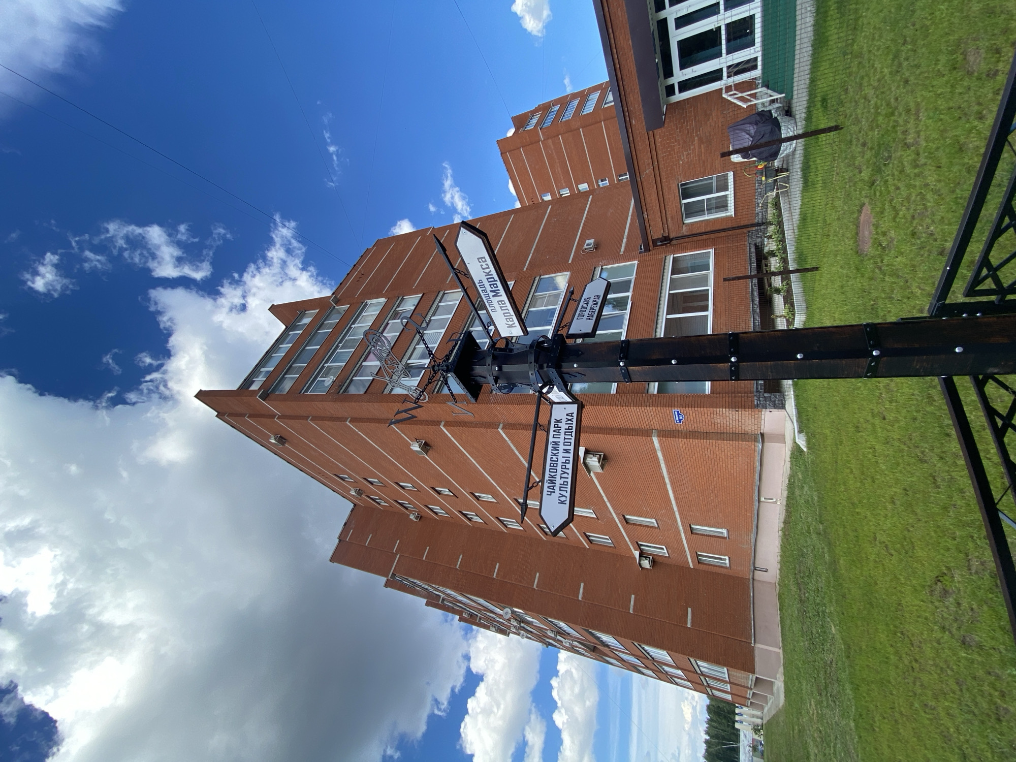 A street signpost stands in front of a modern brick building under a bright blue sky with scattered white clouds. The signpost has multiple white directional signs with black text, indicating locations. One sign reads "ЧАЙКОВСКИЙ ПАРК КУЛЬТУРЫ И ОТДЫХА" which translates to "Chaikovsky Park of Culture and Recreation". Another sign is partially visible, likely indicating another direction. The building appears to be an apartment complex or office building with many windows. The ground in front of the building is covered in neatly trimmed green grass. The scene suggests a pleasant daytime in an urban or suburban setting in Chaikovsky, Russia. The overall mood is serene and ordinary, with no discernible human activity.