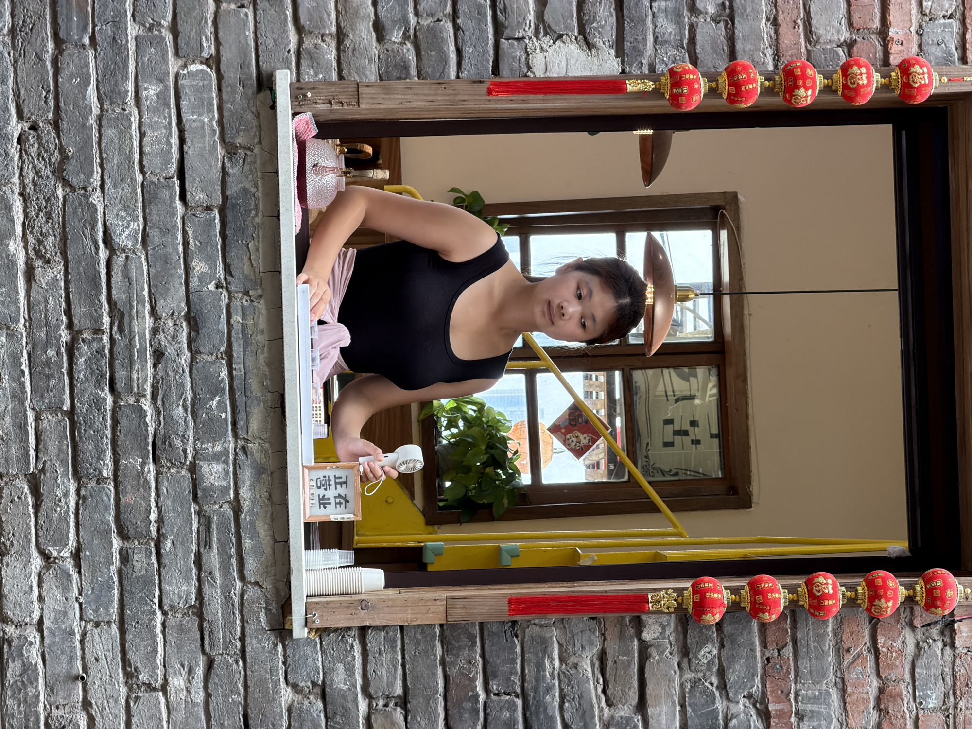 A young East Asian woman with dark hair pulled back stands behind a window in what appears to be a shop or cafe in Yuzhong District, China. She is wearing a black tank top and a light pink sash around her waist. In her right hand, she holds a small white fan. In front of her on the windowsill are several white paper cups and a small framed sign with Chinese characters that read "正在营业" (Zhengzai yingye), meaning "Open for business." To the left, a small glass teapot sits on a pink cloth. The window is framed by rustic brick walls and decorated with strings of red lanterns. Inside the shop, a staircase with a yellow railing and a potted plant are visible. The overall scene suggests a small, traditional business operating in an urban setting.