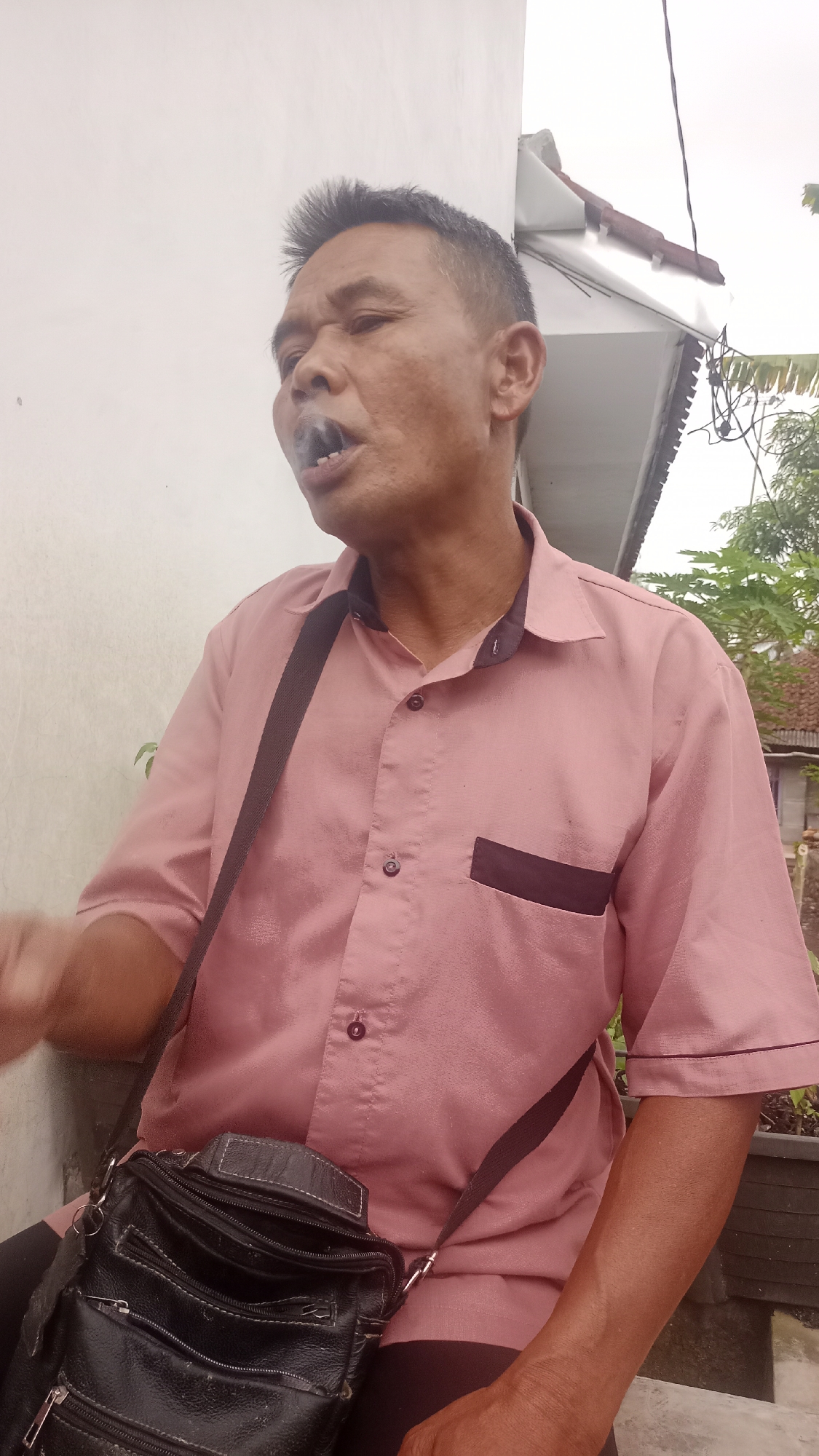 A man is shown outdoors, in what appears to be a residential area. He is wearing a short-sleeved pink button-up shirt with a dark trim on the collar and a dark rectangular patch on the left chest pocket. He has a dark cross-body bag slung over his left shoulder. The man is exhaling smoke, with his mouth open and his eyes looking upwards and to the left. The background includes a white wall, a portion of a roof with terracotta tiles, and some greenery. The setting suggests it is daytime.