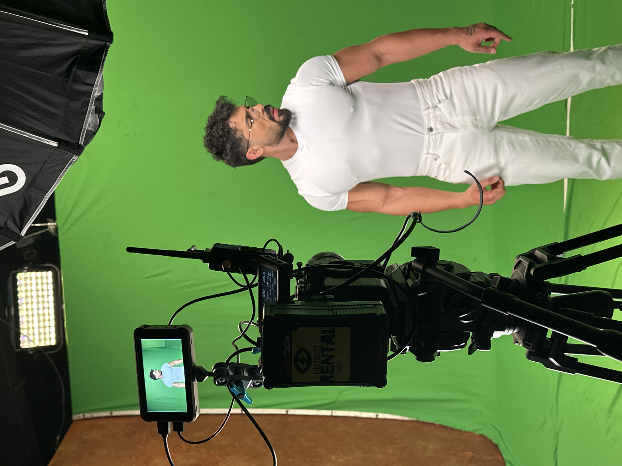 The image depicts a film or video shoot in progress on a green screen stage. A man wearing white clothing is positioned against the green screen. A professional video camera is in the foreground, equipped with a monitor displaying the image being captured. Additional lighting equipment is visible to the left of the frame. The environment appears to be a studio or production space. The location is Sofia, Bulgaria, based on the "Electricity Rental" battery visible on the camera which is located in Sofia.