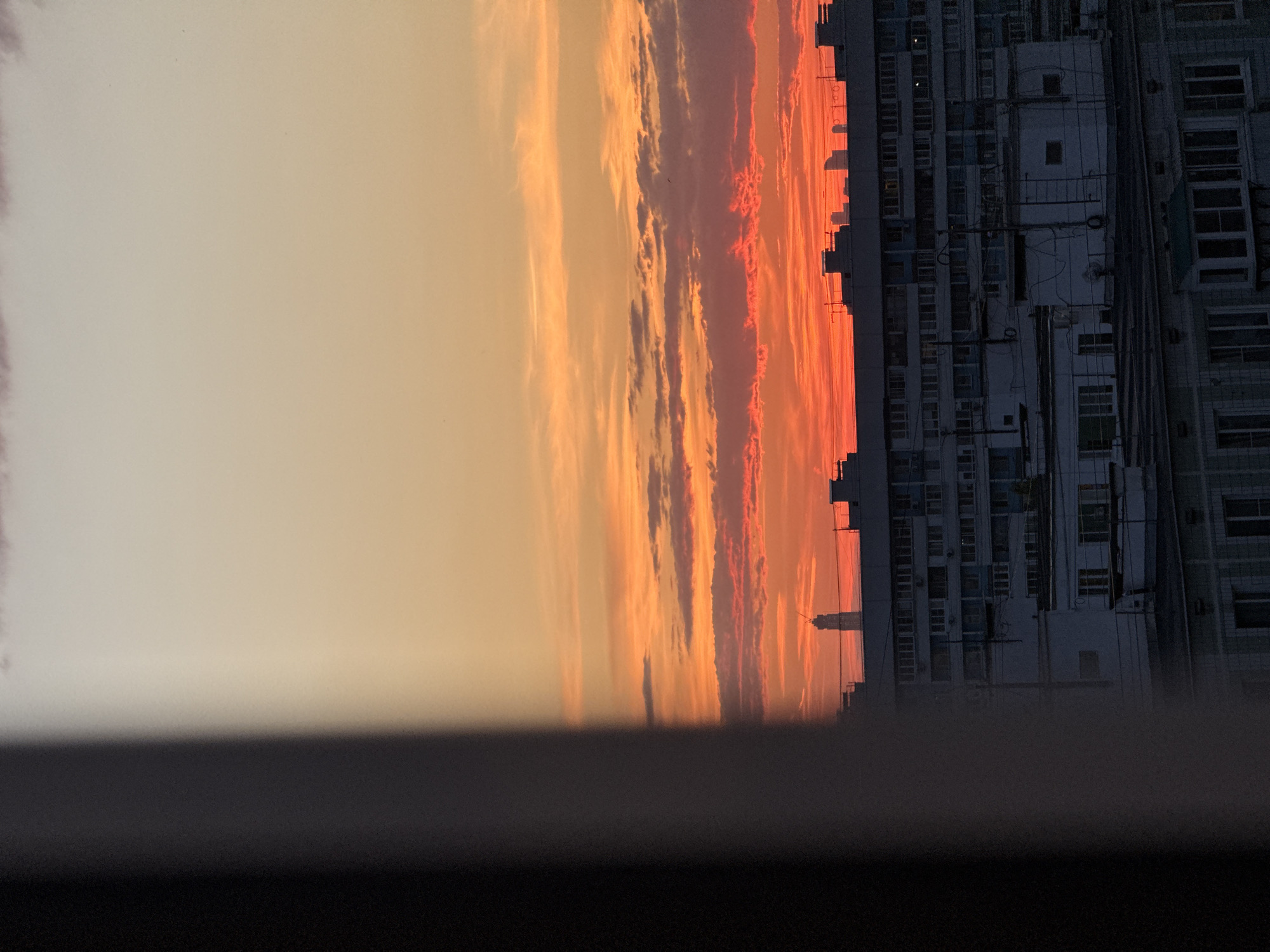 This image captures a dramatic urban sunset or sunrise over Moscow, Russia, viewed from an elevated perspective, possibly a window or balcony. The sky dominates the left and upper portions of the frame, presenting a spectacular display of colors. Bands of clouds stretch horizontally across the sky, transitioning from a soft, pale orange and yellow on the far left to vibrant, fiery reds and deep oranges closer to the center and right. The light source appears to be to the right, illuminating the edges of the clouds and creating a stark contrast.
On the right side of the image, a dense cityscape of multi-story buildings is silhouetted against the intensely colored sky. These structures, likely apartment or office blocks common in urban Moscow, appear dark grey to black due to the backlighting. Numerous windows are visible, some reflecting faint light, others dark recesses. Architectural details like balconies, air conditioning units, and utility lines can be discerned on the facades. A distinct, taller building with a noticeable spire or antenna-like top stands out against the horizon among the other buildings, hinting at a landmark or a particularly high-rise structure within the urban sprawl.
The bottom portion of the image features a dark, blurry horizontal band, suggesting a window frame or balcony railing, grounding the viewer's perspective. The overall scene evokes a sense of awe and tranquility, showcasing the beauty of natural light against a bustling metropolitan backdrop at either dawn or dusk. There is no visible text in the image.