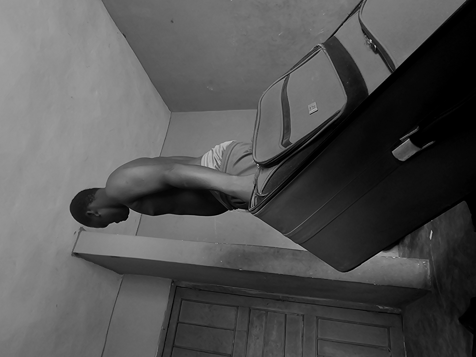 A black and white photograph shows a shirtless man from the waist up, facing away from the camera, holding the handle of a dark suitcase. He is in what appears to be an indoor space. To his left, a wooden door is visible. The room has concrete walls and a textured ceiling. The man is wearing light-colored shorts. The suitcase is large and appears to be a piece of luggage. The lighting suggests an indoor environment, possibly a home or a lodging. The specific location in Nsugpe, Nigeria, is not discernible from the image itself.