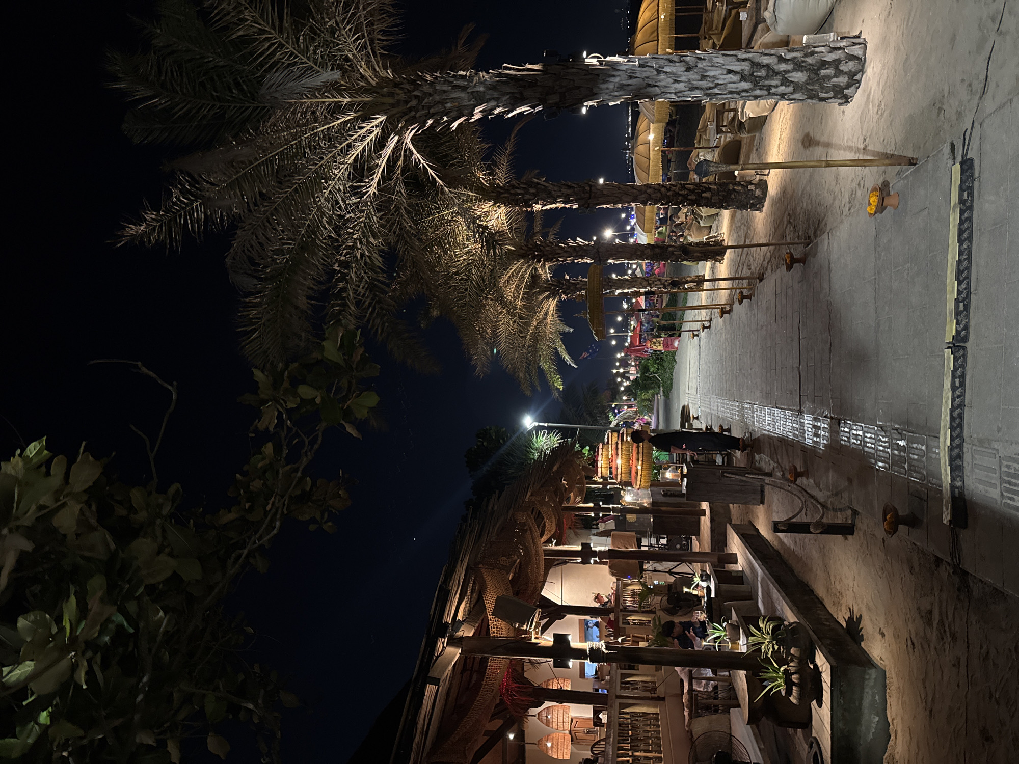 This image depicts a beachfront restaurant or lounge area at night in Kuta, Indonesia. The scene is illuminated by ambient lighting from within the establishment, strategically placed torches, and string lights visible in the background.
On the left, a restaurant with a thatched roof and open-air seating is visible. The interior is dimly lit, revealing tables, chairs, and patrons. Decorations include fishing nets draped over the roof structure and hanging lanterns. Plants in pots are placed on wooden platforms.
A paved walkway runs alongside the restaurant, separating it from the sandy beach area. Palm trees line the walkway, with some illuminated from below, casting shadows. Several beach umbrellas and low seating areas with beanbags are set up on the sand to the right of the walkway.
A few individuals are present, including staff members and customers. One staff member is standing near the walkway, and another appears to be serving guests inside the restaurant. In the background, more beachgoers are visible. The night sky is dark, with faint stars. The overall atmosphere suggests a relaxed evening setting at a tropical resort.