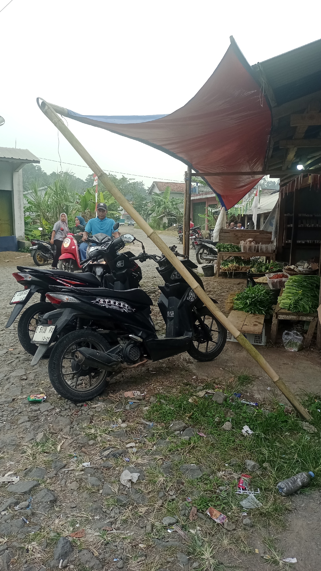 The image depicts a roadside market scene in Karyamandala, Indonesia. In the foreground, two black motorbikes are parked on a gravel and dirt surface. The motorbike on the left has a license plate that reads "B 5138 RX 12-28". The motorbike on the right has "STREET" written on its side panel.
Behind the motorbikes, a red motorbike is visible with two people on it. The person in front, a man wearing a blue shirt and a cap, appears to be looking forward. The person behind him is wearing a green headscarf. Further to the left, another woman wearing a grey headscarf is standing next to a black motorbike.
A makeshift canopy, made of red and blue tarpaulin supported by a large bamboo pole, is erected over a market stall. The stall displays various fresh produce, including leafy greens, red chilies, and other vegetables. The background shows houses and vegetation, suggesting a semi-rural or suburban setting within Karyamandala. The ground is littered with some debris, including plastic wrappers and a water bottle. The sky is overcast.