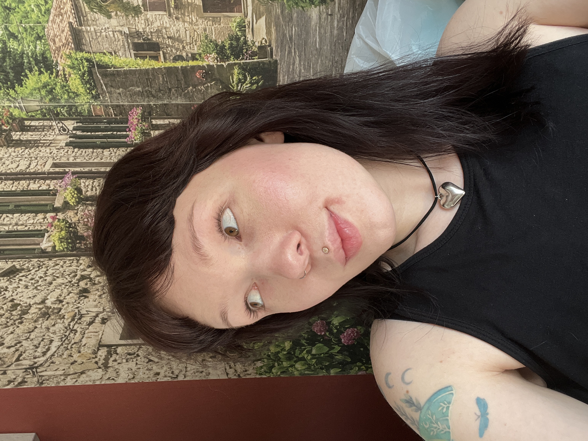 A young woman with brown hair and fair skin is pictured. She wears a black tank top and a silver heart necklace. She has a lip piercing on the right side of her upper lip. She also has tattoos on her upper left arm.
The background appears to be a patterned wallpaper or backdrop depicting an outdoor scene of buildings and greenery, possibly meant to resemble a European cityscape or countryside scene.
The location, Chaikovsky, Russia, is not visually evident from the provided image. There are no identifiable landmarks or cultural elements to indicate the location.