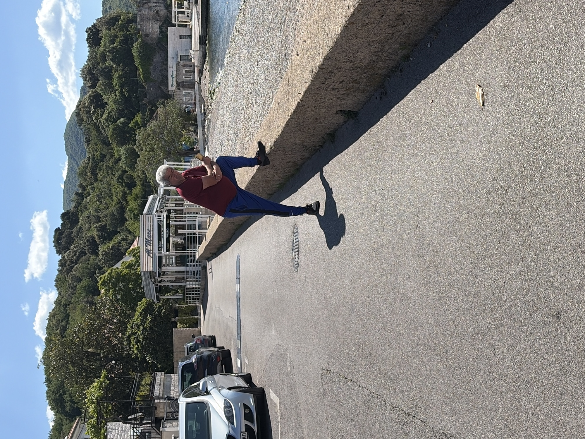 An eye-level shot reveals a man sitting on a concrete wall, with his back against the wall and legs extended out onto the asphalt. He wears a maroon shirt and blue pants. In the background, trees and a building with "Al Mare" in large lettering are visible. The asphalt is cracked and leads to parked cars in the distance. A single leaf lies on the asphalt. The location is likely in Meljine, Montenegro, given the building signage.