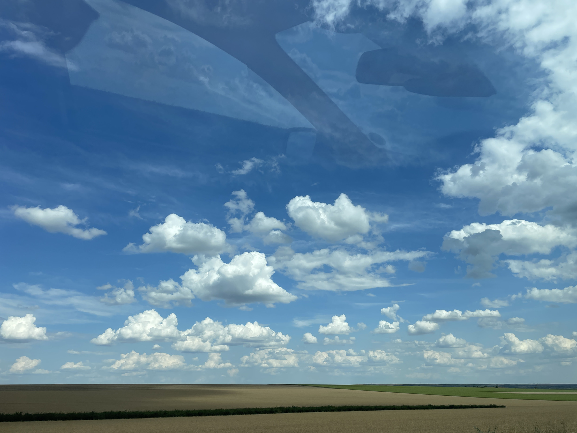 The image depicts a vast, flat landscape under a vibrant blue sky filled with numerous white, puffy cumulus clouds. The foreground shows agricultural fields, primarily in shades of brown and gold, with a thin dark green strip of vegetation along the edge of the closest field. In the distance, the landscape appears to continue with more fields, some of which are a brighter green, suggesting different crops or stages of growth. The sky is clear and expansive, with some darker, more diffuse cloud formations visible in the upper part of the image, possibly reflections on a surface. The overall scene is one of rural tranquility and wide-open spaces, characteristic of agricultural regions. There are no people or specific objects visible that would indicate an urban setting within Comrat, Moldova. The image appears to be taken from a slightly elevated perspective, possibly from a vehicle or a gentle rise in the terrain.