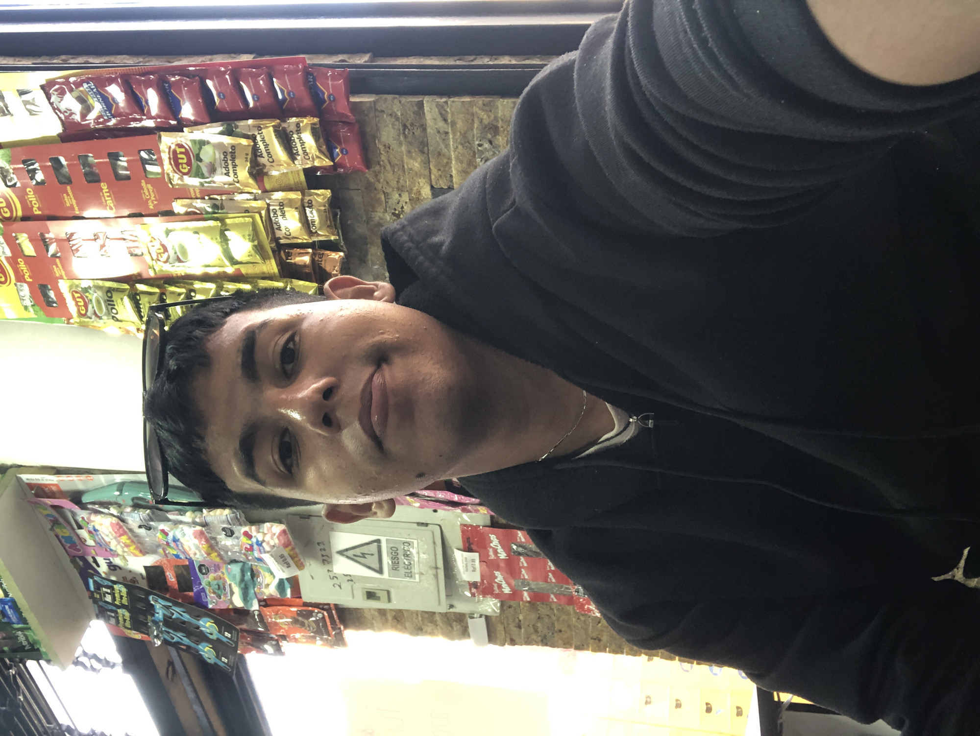 The image shows a young person wearing a black hoodie, taking a selfie in a store. The background features shelves stocked with various packaged goods, including what appear to be spices and candies. A prominent sign with a lightning bolt symbol and the words "RIESGO ELECTRICO" (Electric Risk) is visible on a grey electrical box on the wall. The store's ambiance suggests a small neighborhood shop or bodega. The person is looking directly at the camera with a neutral expression. The lighting suggests an indoor environment, possibly during the day given the brightness from a window or overhead lights. The setting appears to be a commercial establishment in Juan Griego, Venezuela, a coastal town known for its port and historical sites.