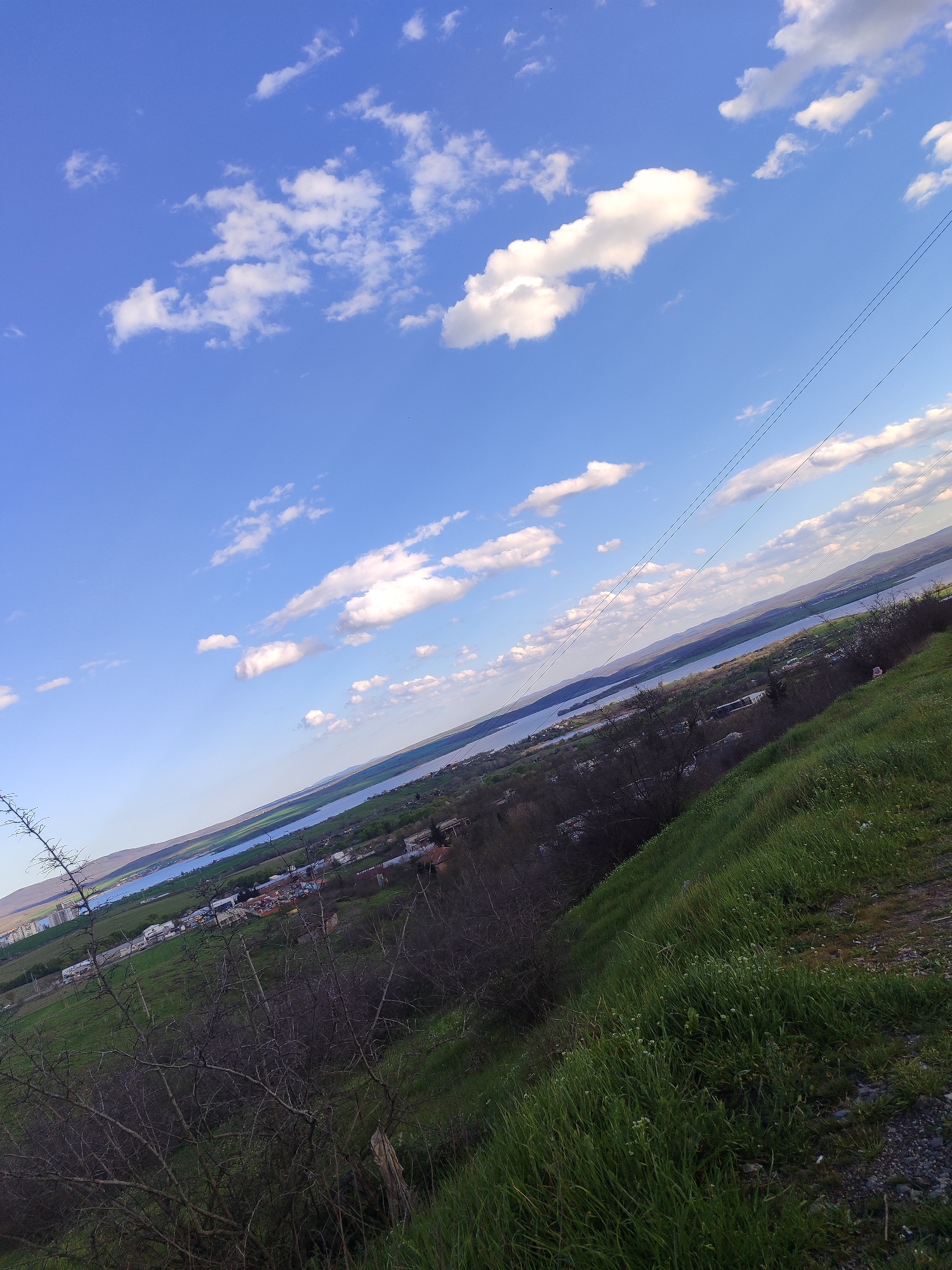The image shows a landscape view with a blue sky and white clouds taking up the majority of the frame. The land slopes downward from the bottom right corner towards the middle. In the distance, there is a body of water and a cityscape that possibly indicates the city of Burgas, Bulgaria. A few trees are visible in the foreground on the left side of the image. The overall perspective suggests the image was taken from a high vantage point. Power lines are visible diagonally across the sky from right to left.